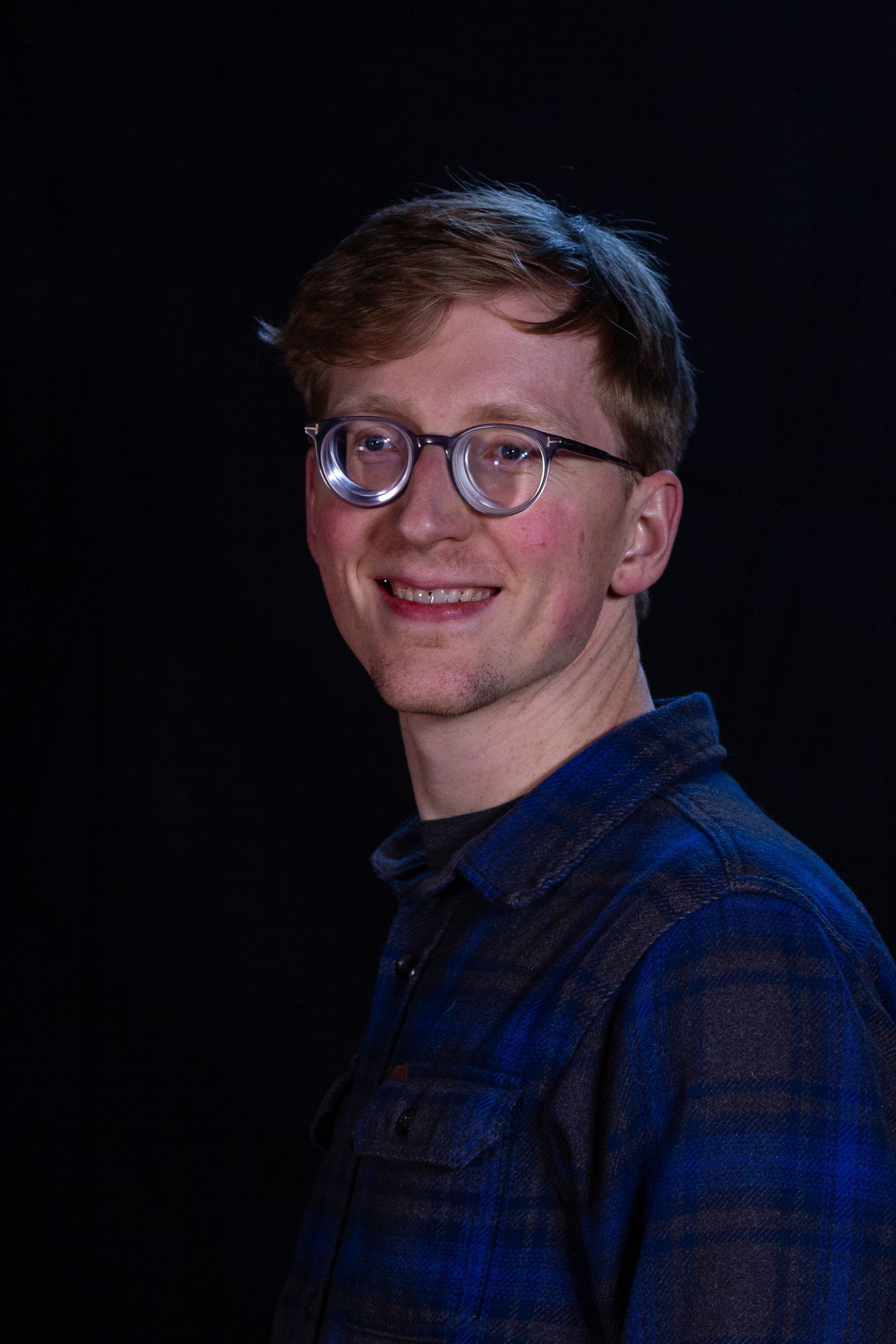 Young man with glasses smiling, wearing a blue plaid shirt, against a black background.