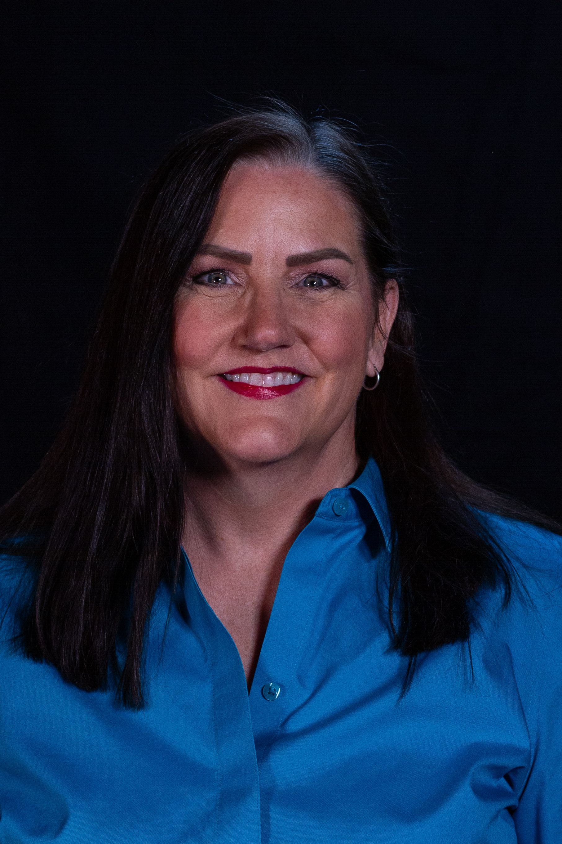 Woman with long dark hair and a blue shirt smiles, looking forward, against a black background.