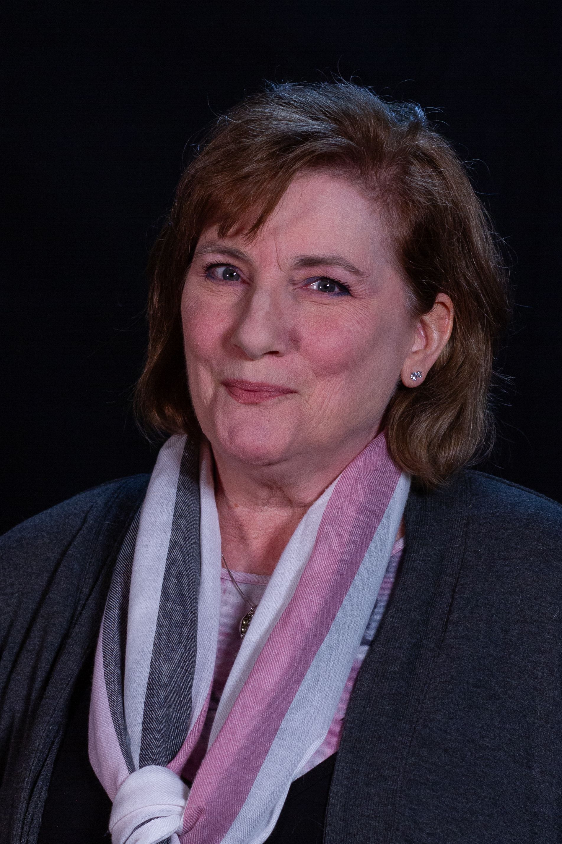 Woman with red hair, wearing a scarf and jacket, smiles at the camera against a dark background.
