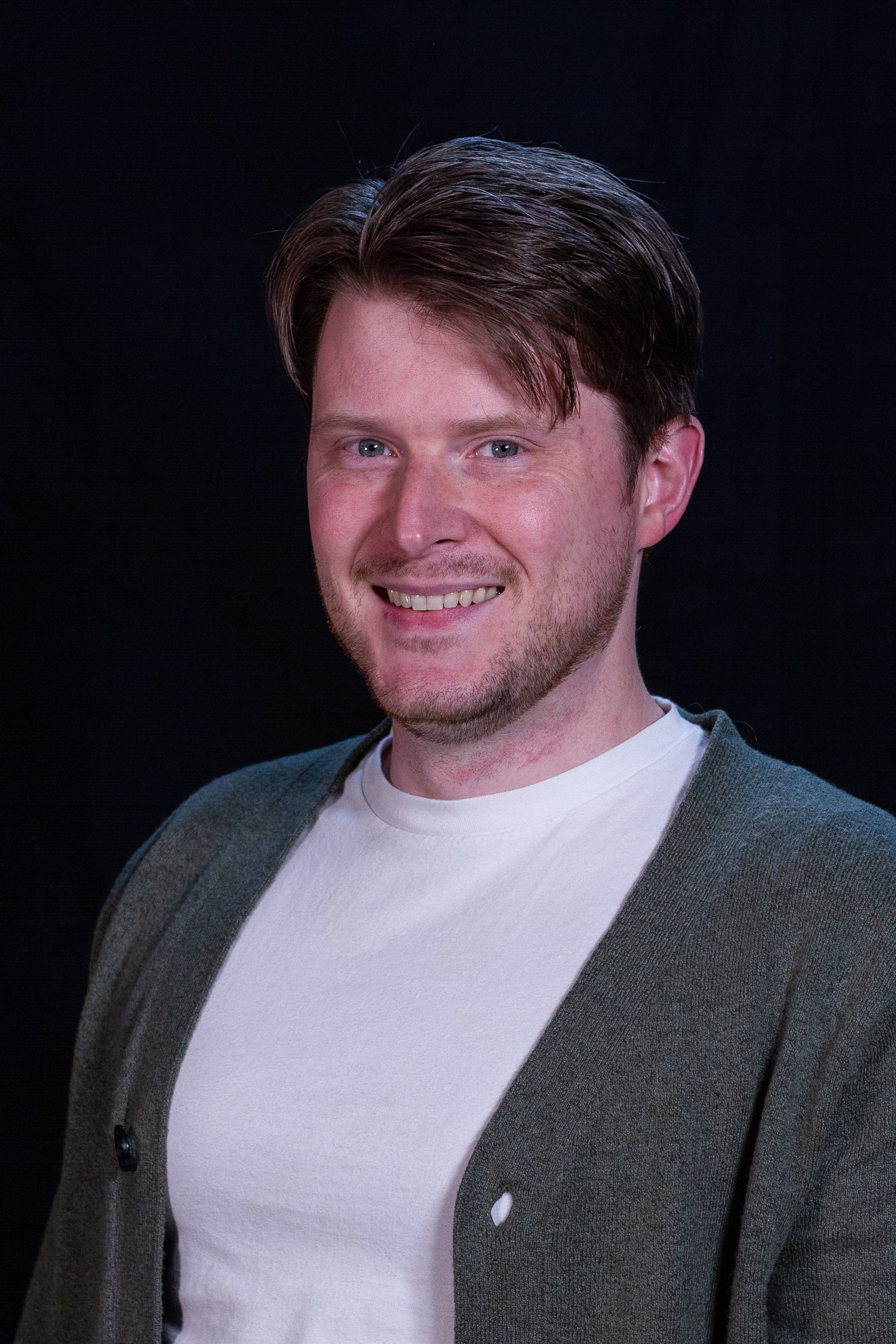 Man with fair skin, smiling, wearing a white shirt and green cardigan against a dark background.