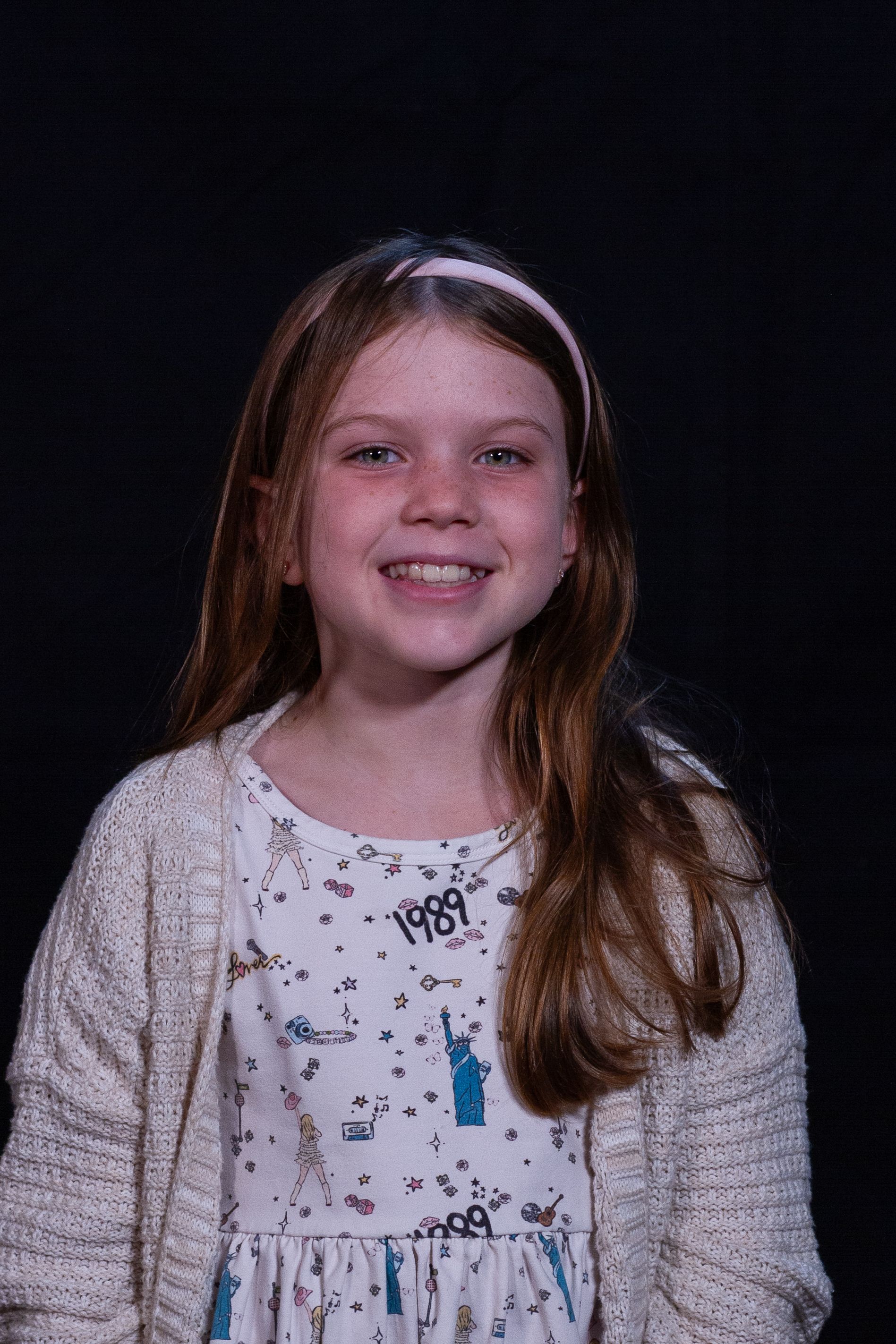 Smiling girl with long brown hair wearing a white sweater and shirt, pink headband, against a dark background.