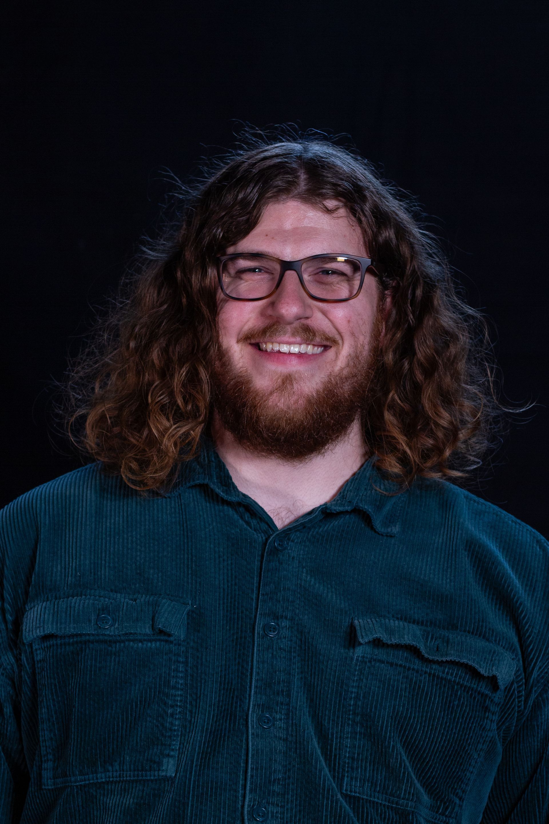 Man with glasses, long curly hair, and beard smiling, wearing a teal corduroy shirt against a dark background.