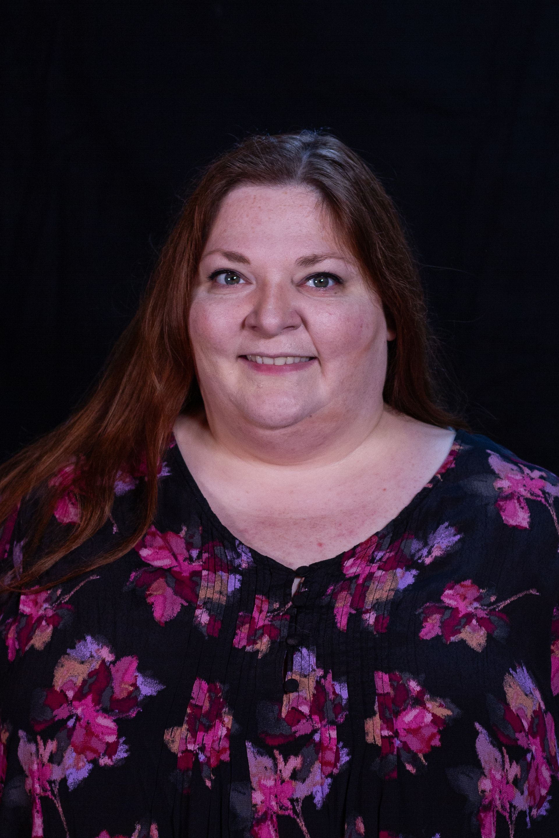 Woman with reddish-brown hair, smiling, wearing a floral top against a black backdrop.