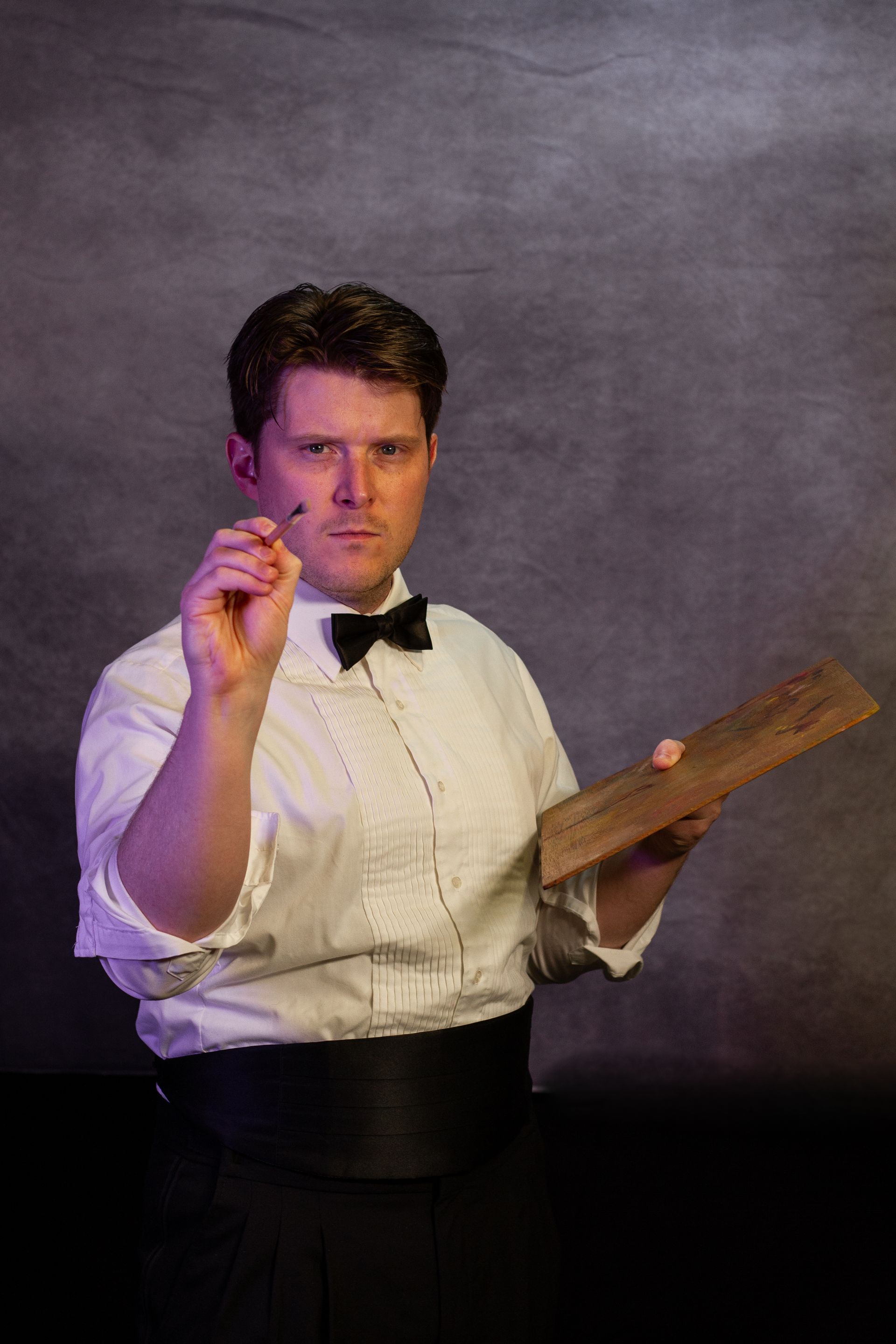 Man in formal wear, holding a pencil and a notepad, pointing, serious expression, against a grey background.