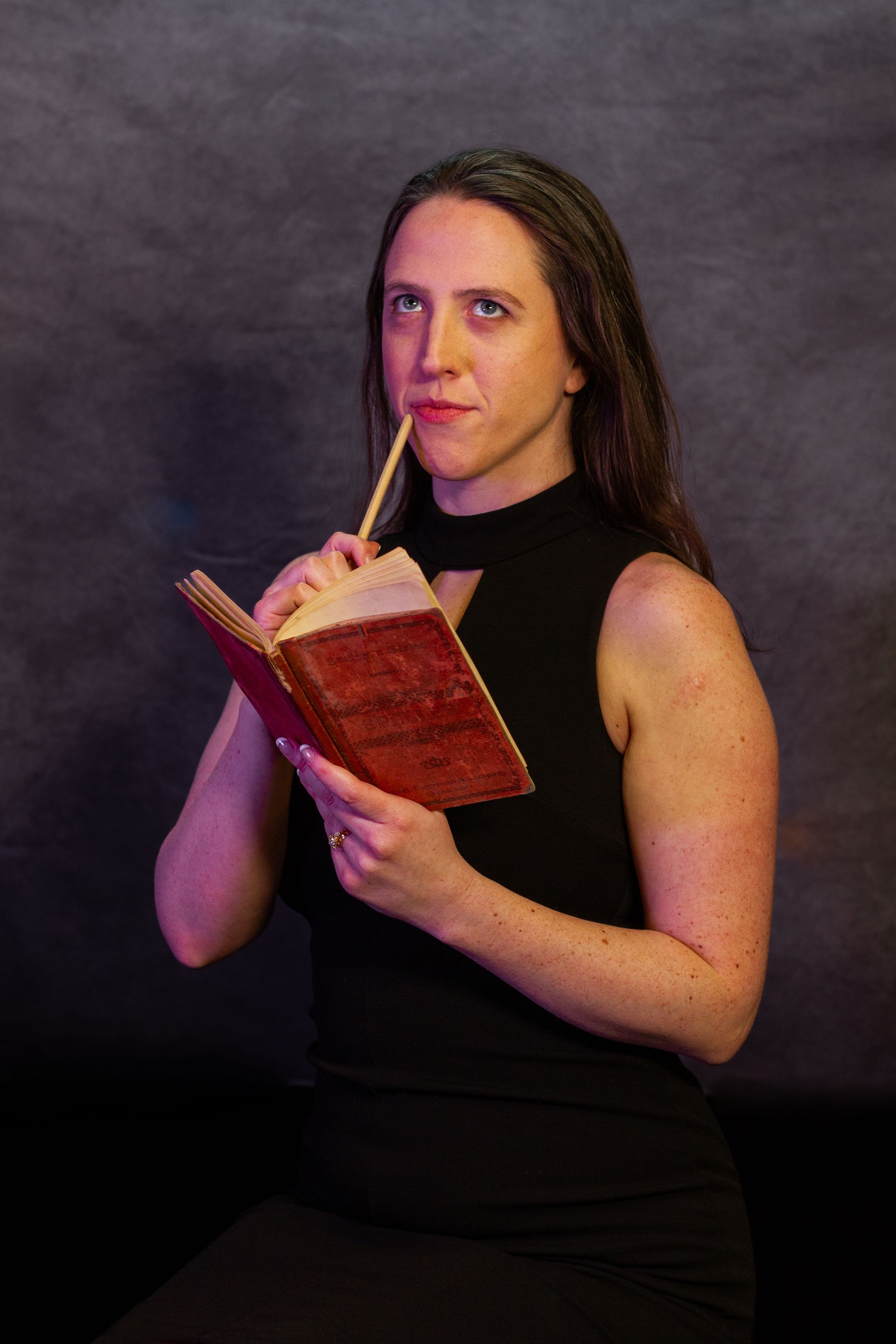 Woman with pen to lips, looking up, holding open book; seated in front of a dark background.