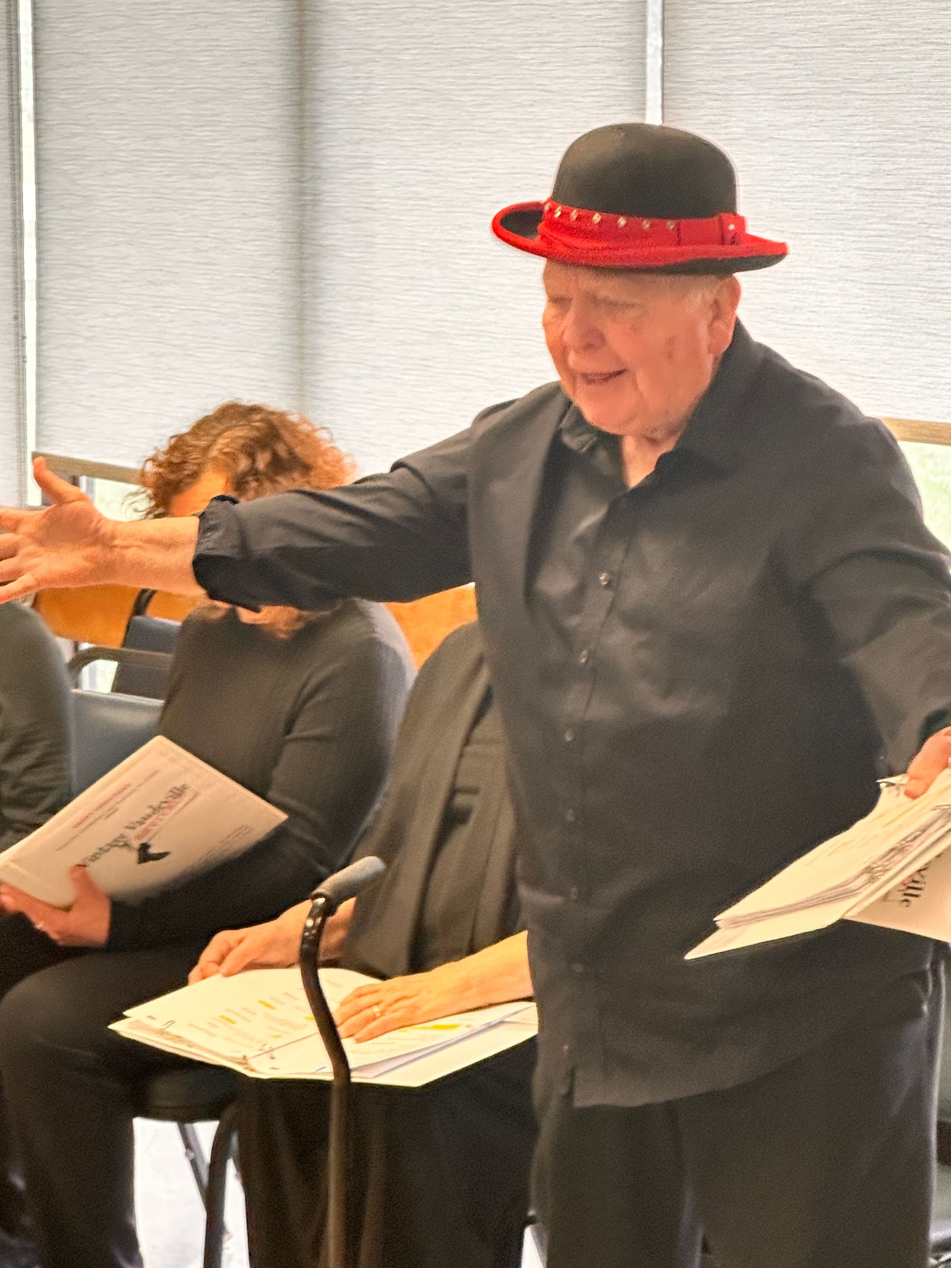 Man in red-trimmed hat gestures with sheet music in a music rehearsal, others seated nearby.