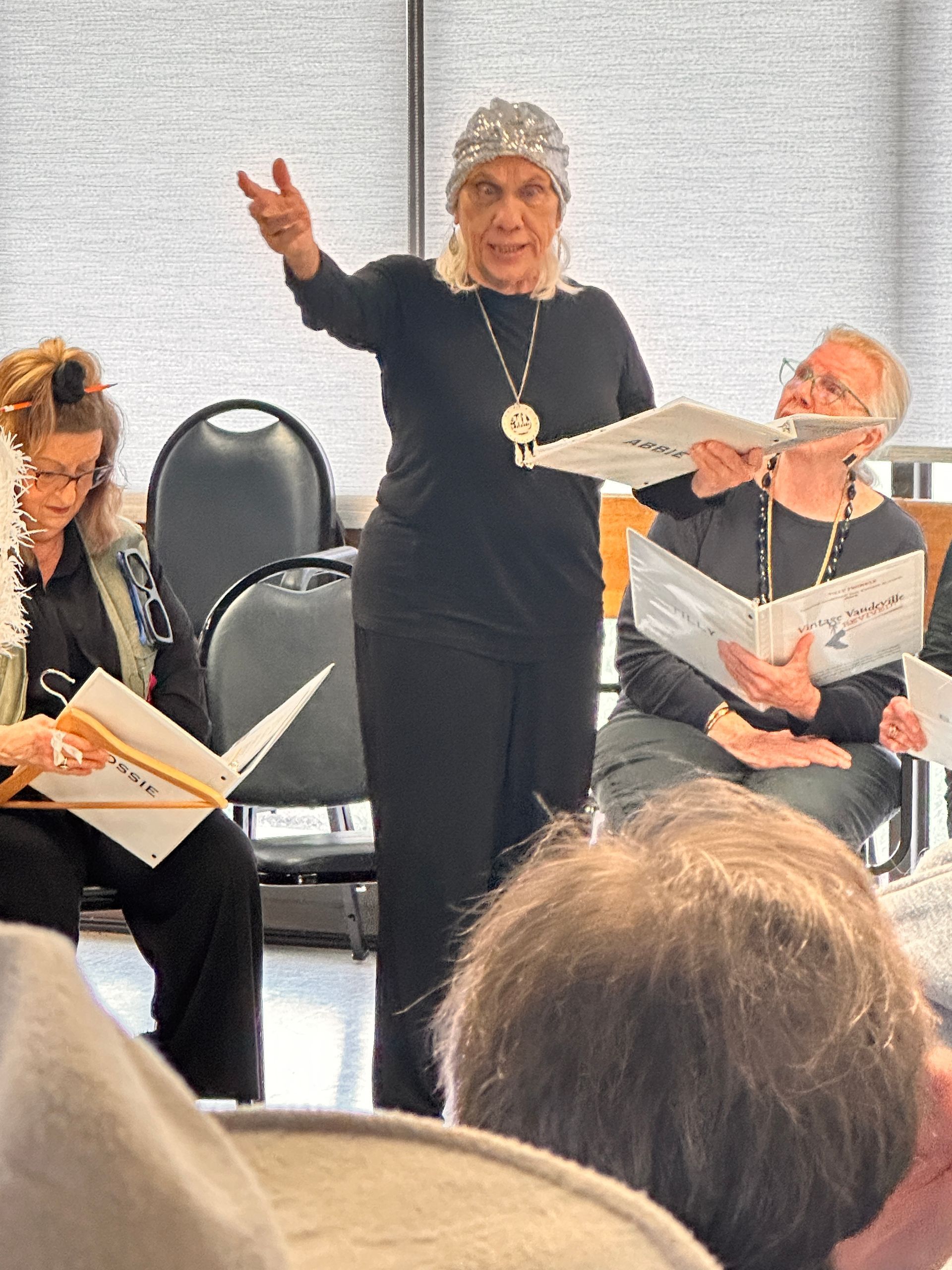 Choir rehearsal: Person with a headscarf conducts, two others hold sheet music, facing audience in a room.