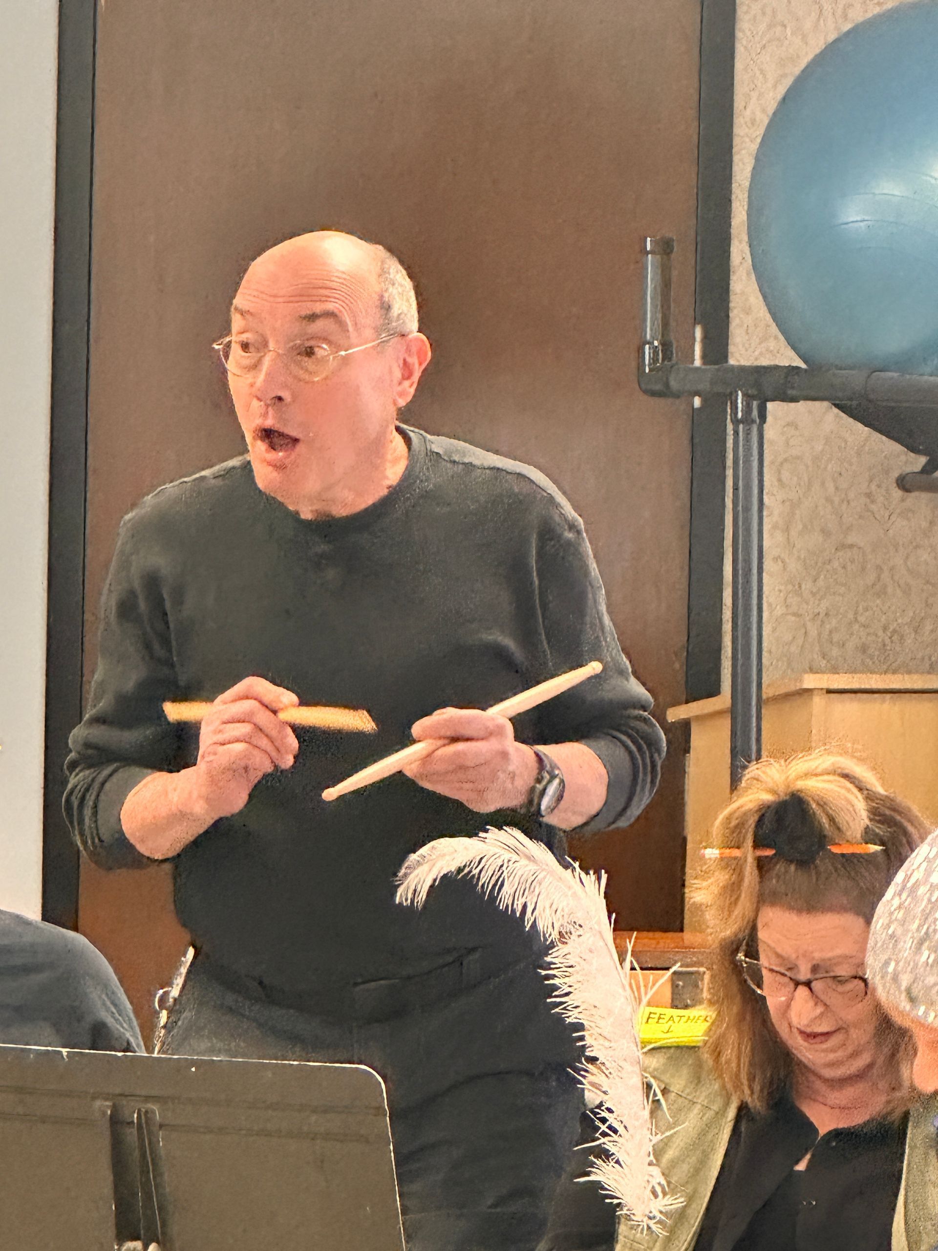 Man with surprised expression, holding broken drumstick. Indoor setting, with a band and musical equipment.
