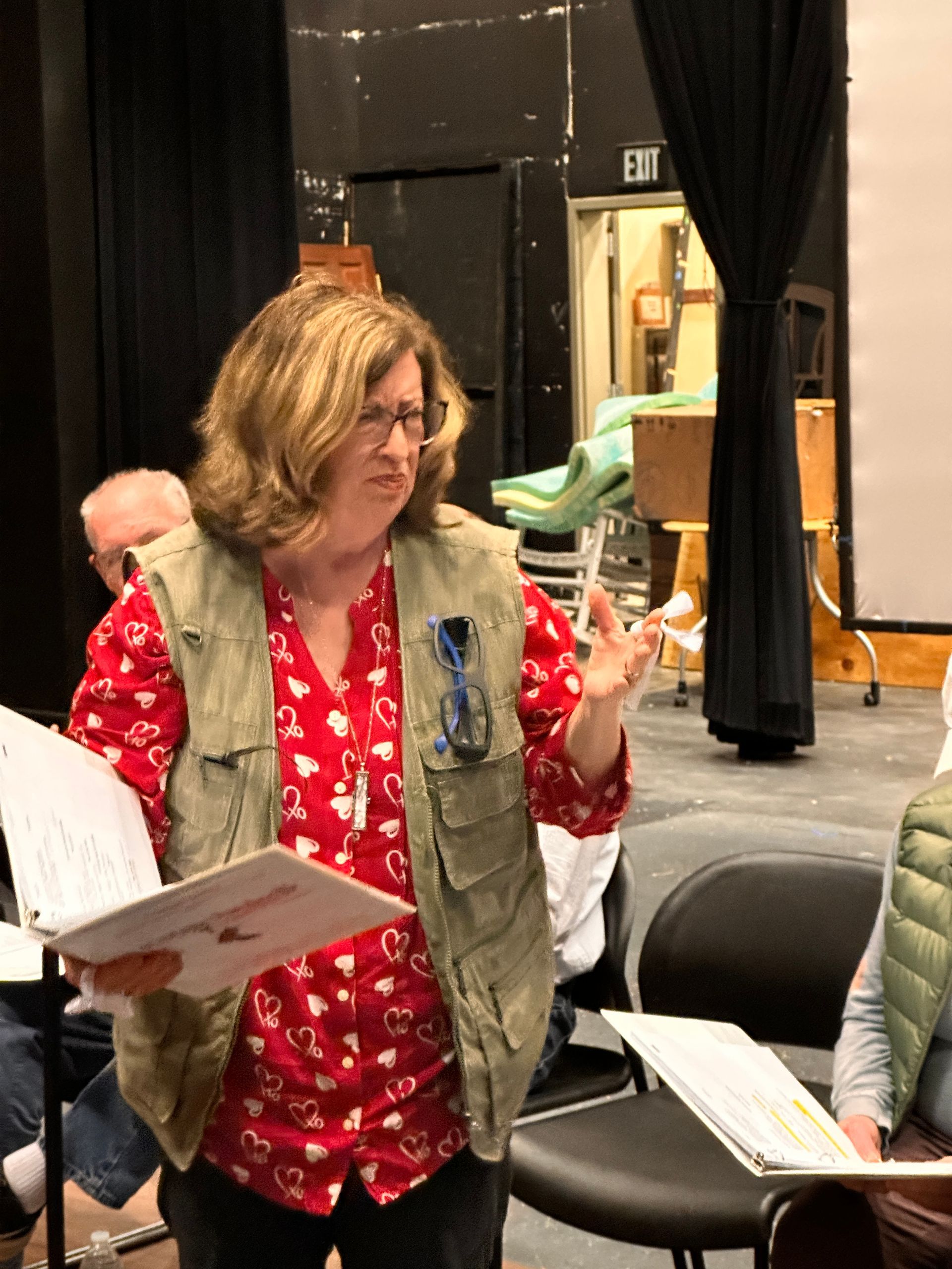 Woman in vest and red shirt gestures while holding music, in auditorium.