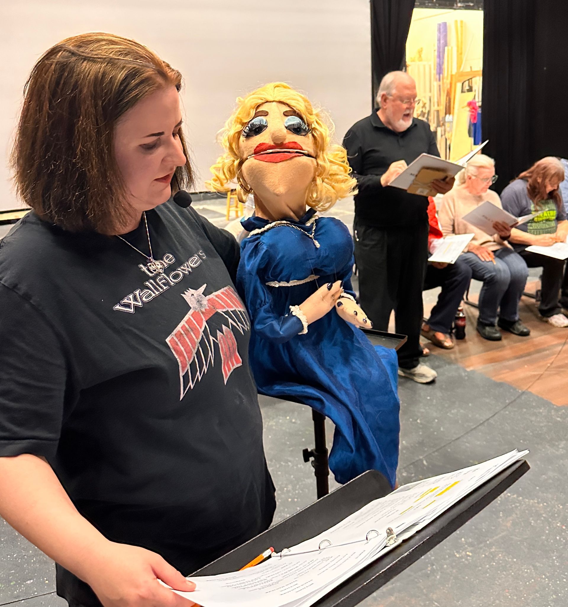 Woman holds puppet while reading script with group. Blonde puppet in blue dress. People seated, reading.
