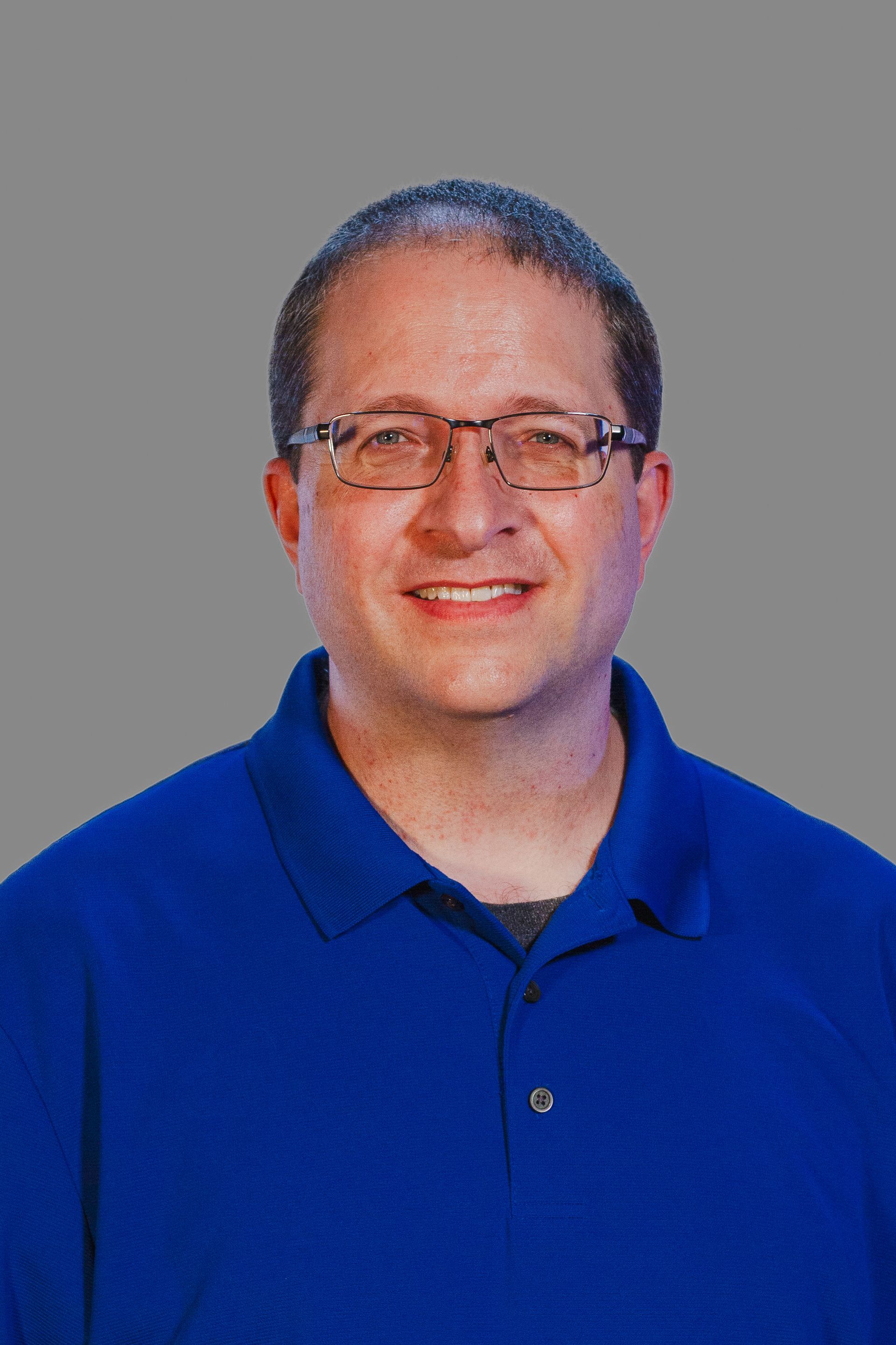 Man with glasses wearing a blue shirt smiles at the camera against a gray background.