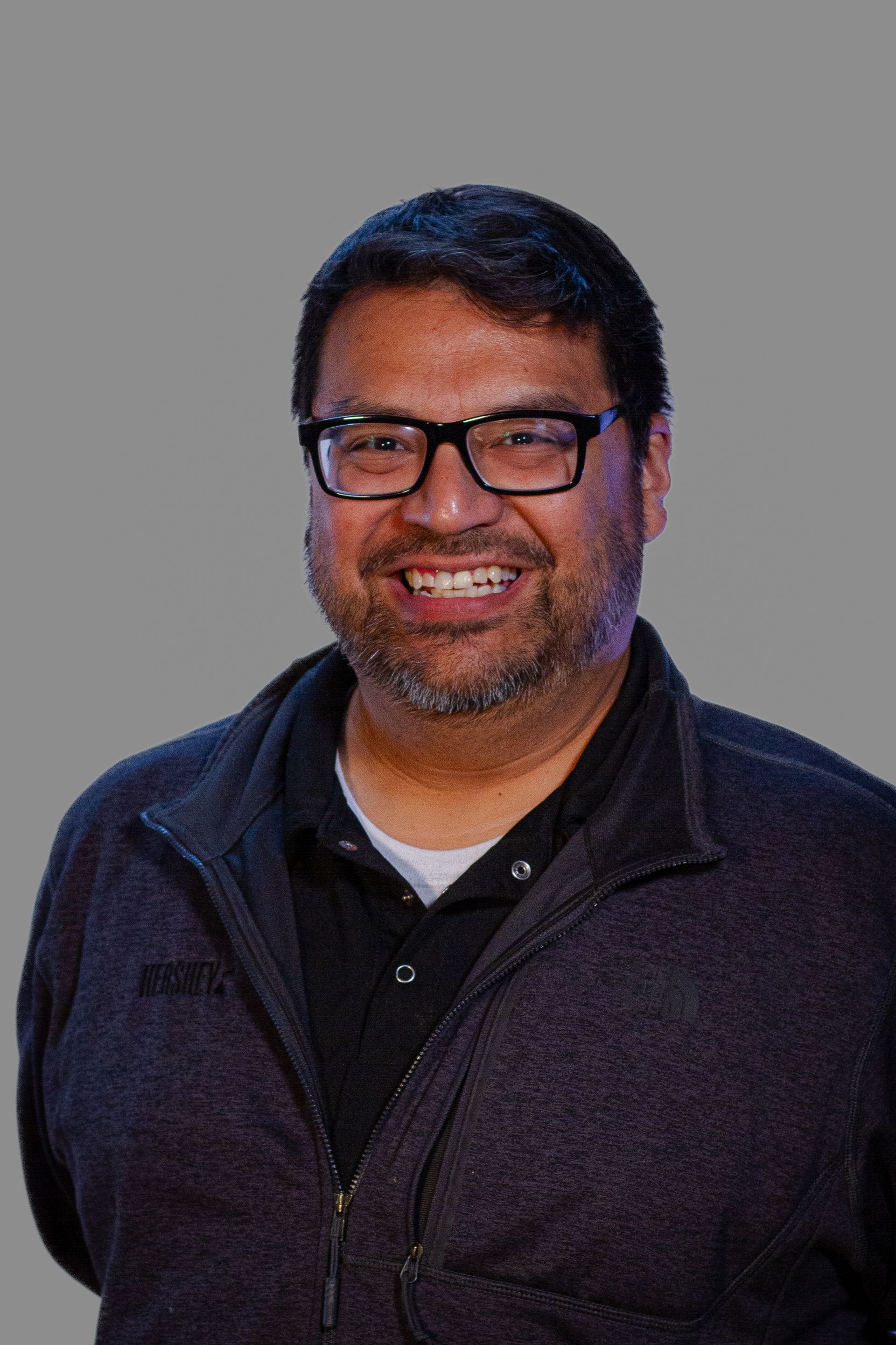 Man wearing glasses smiles, wearing dark jacket over black shirt, against a gray background.