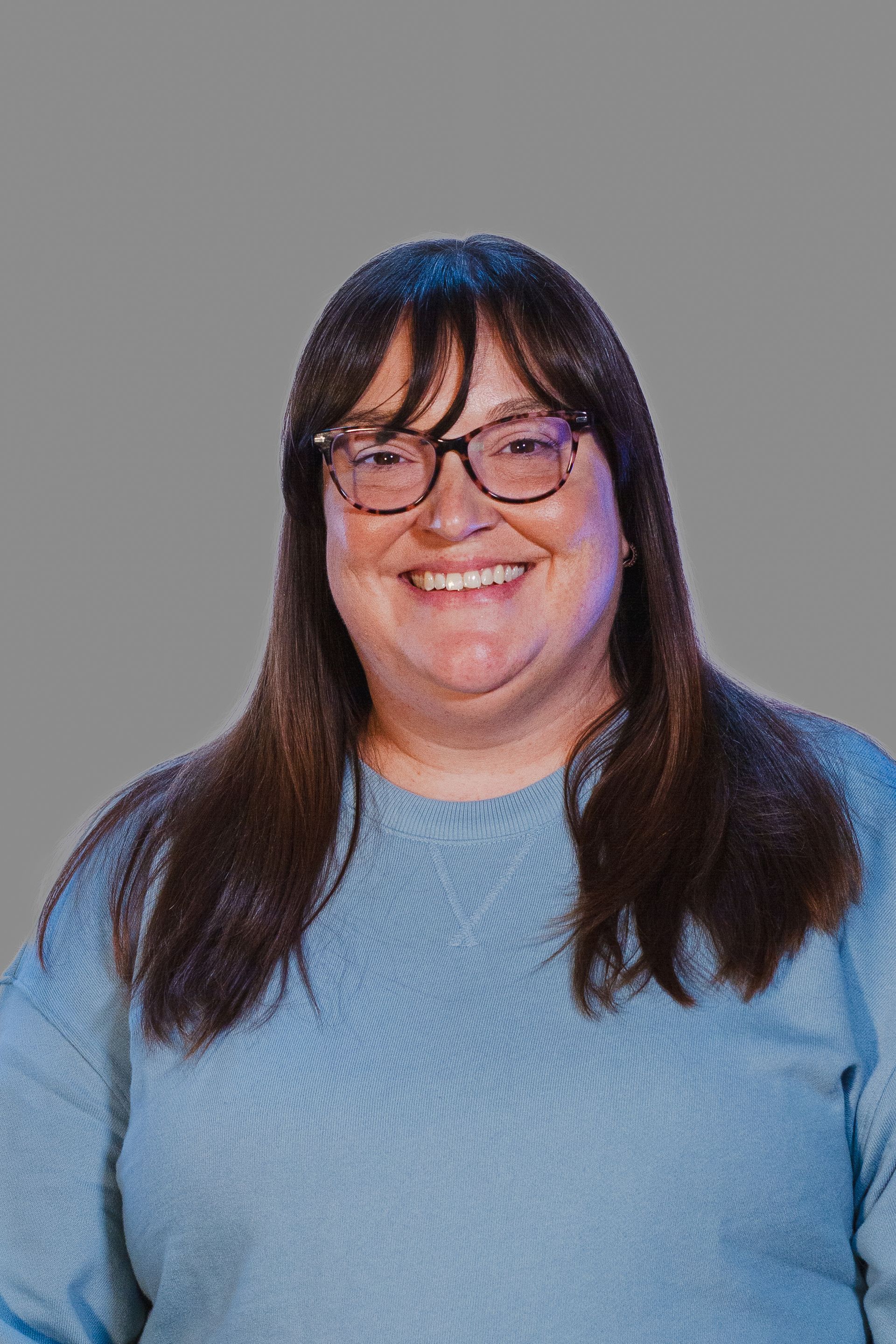 Woman with glasses smiles, wearing a blue sweater, against a gray background.