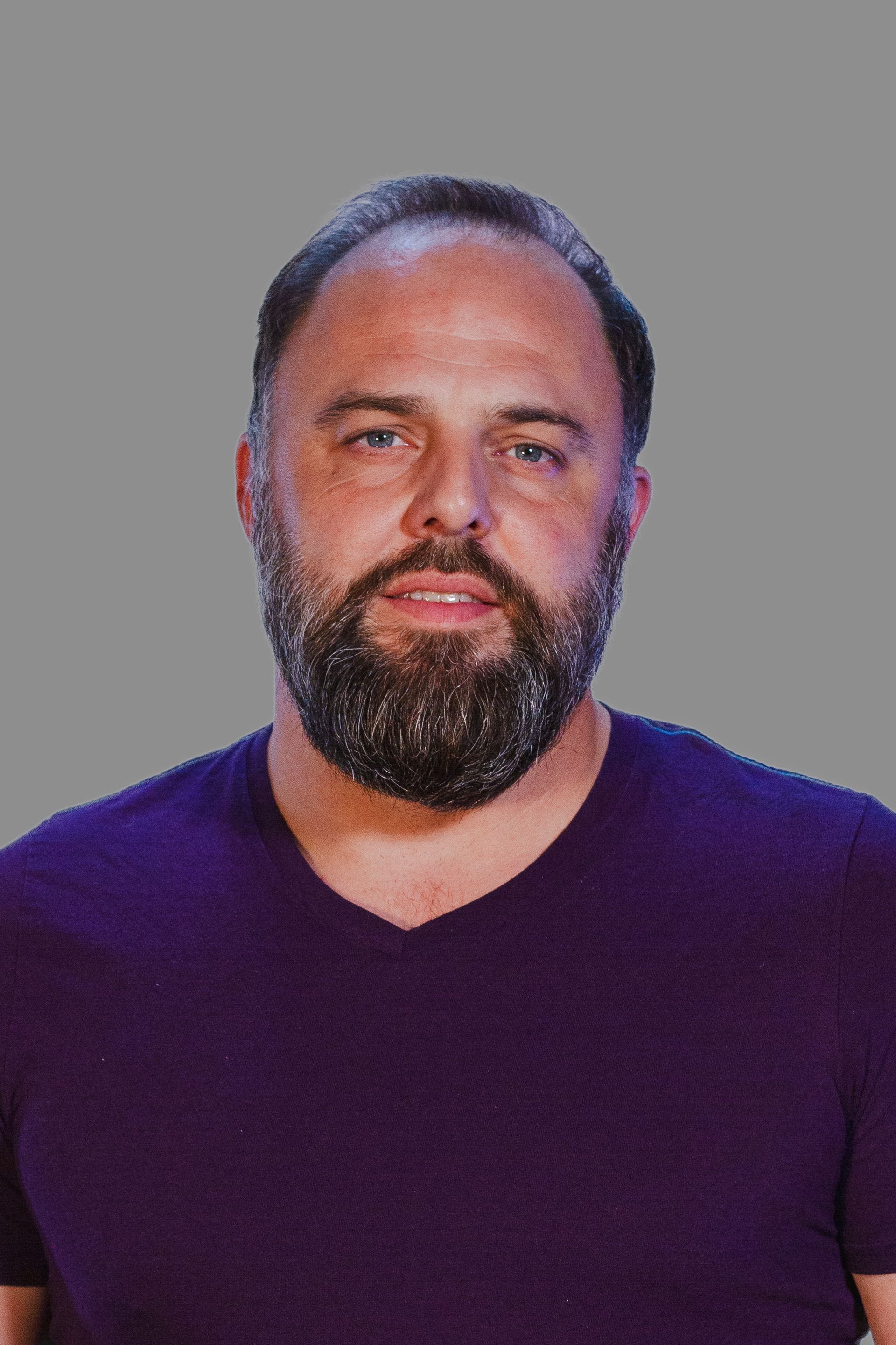Man with a full beard, wearing a purple v-neck shirt, looking at the camera.