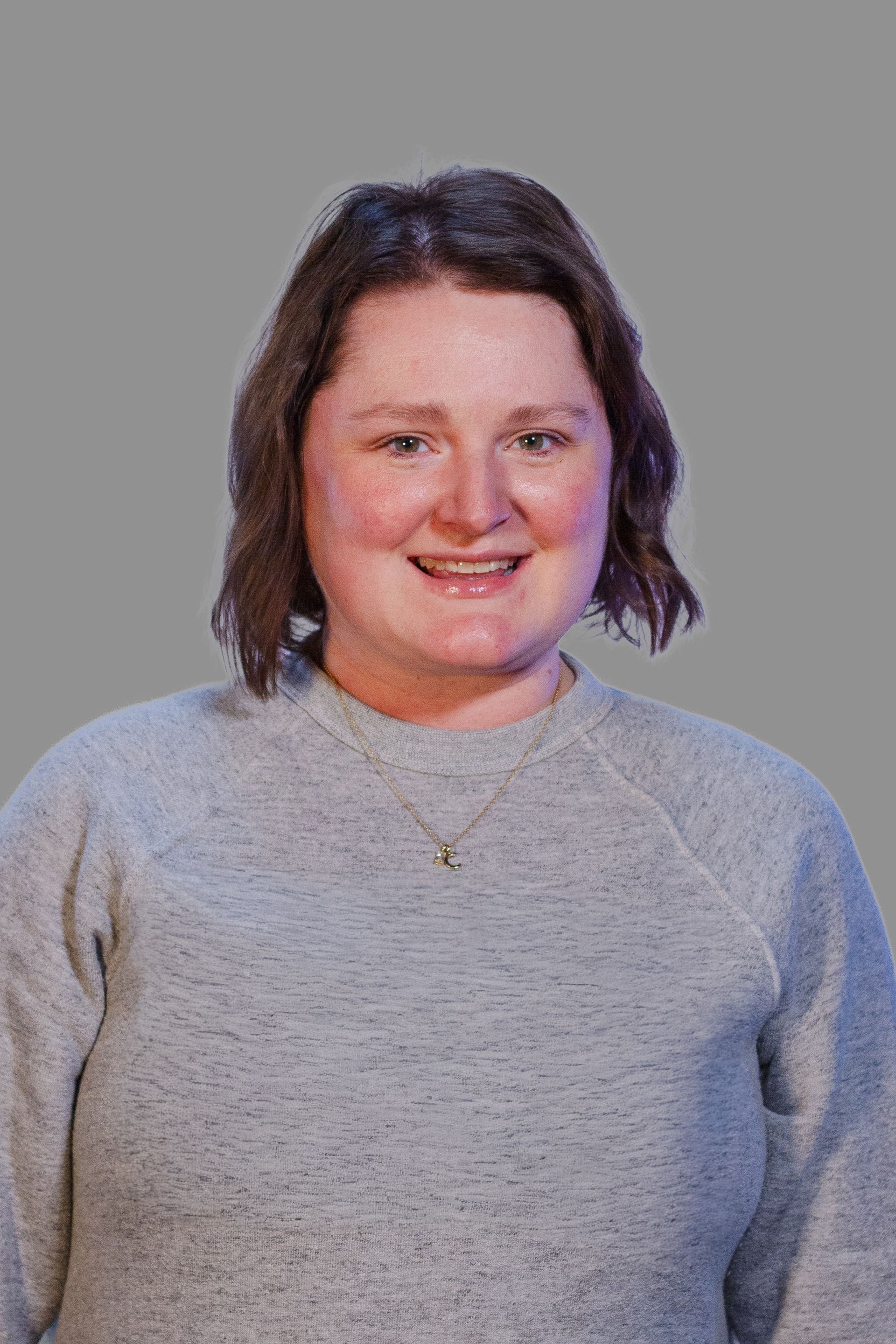 Woman smiling in a grey sweater; light skin, short brown hair, small necklace. Gray background.