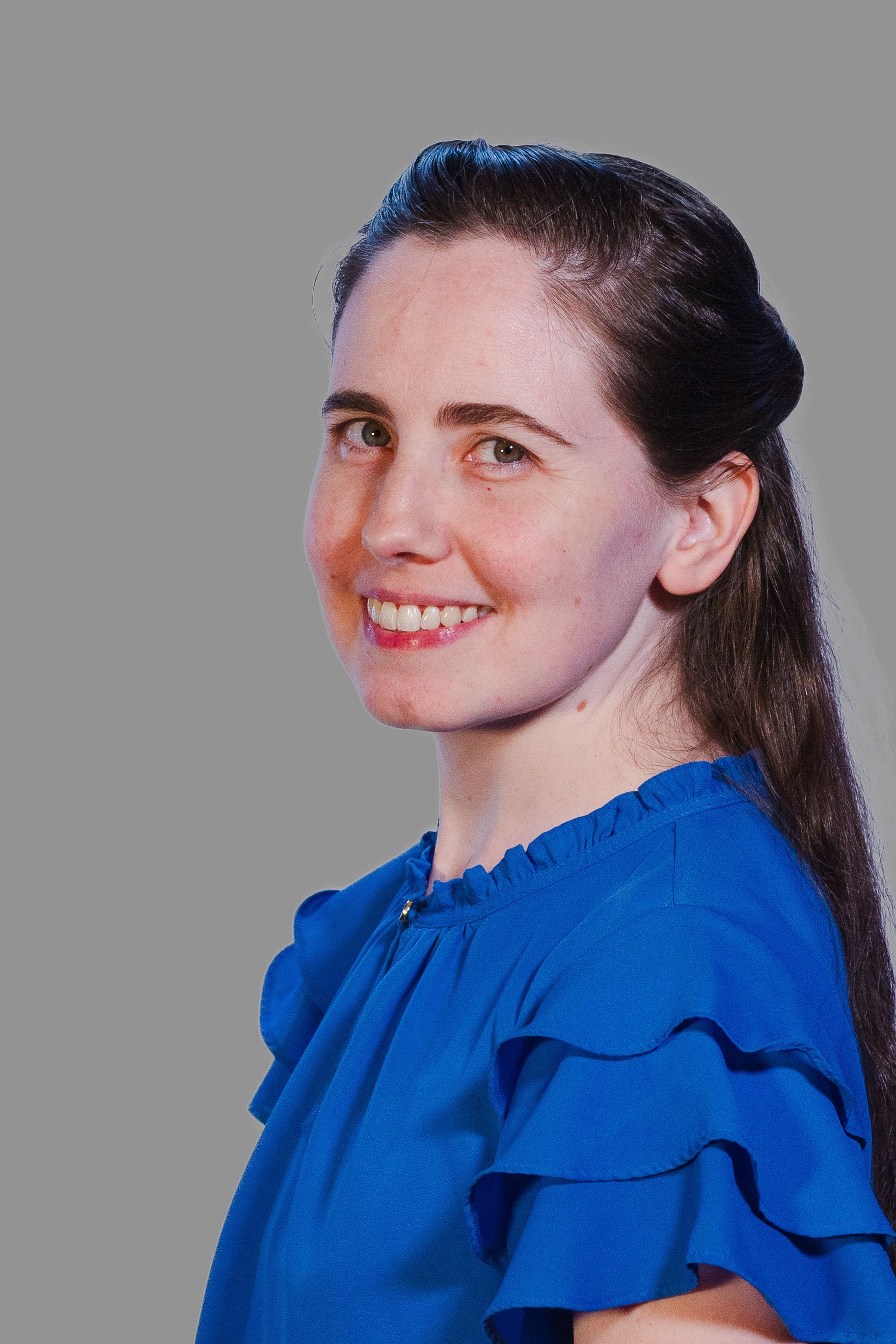 Woman with dark hair, smiling, wearing a blue ruffled shirt, against a gray background.