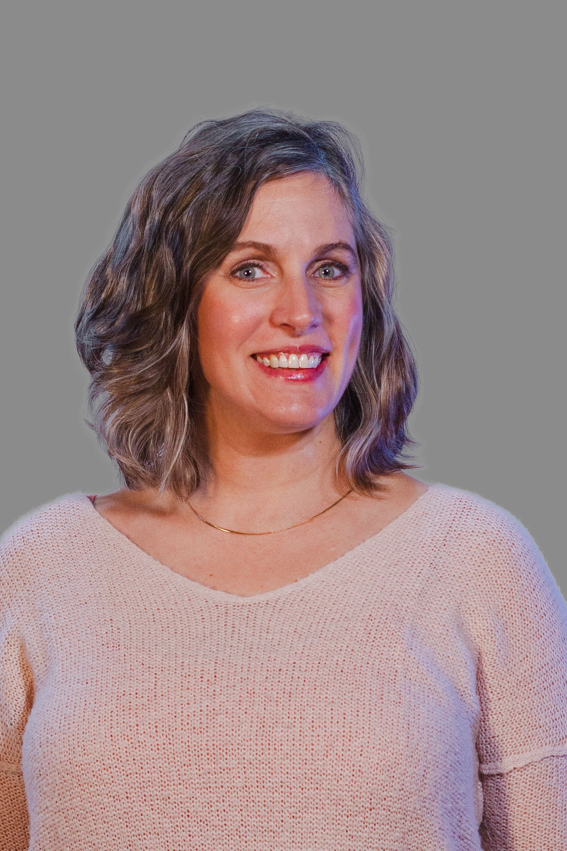 Woman with curly grey hair smiles, wearing a light pink sweater, against a grey background.