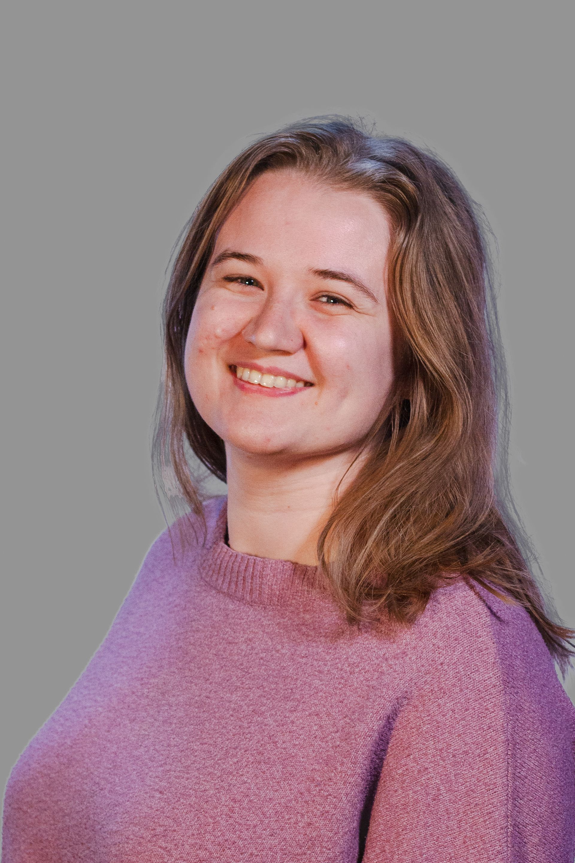 Woman with long brown hair, smiling, wearing a light purple sweater against a gray background.