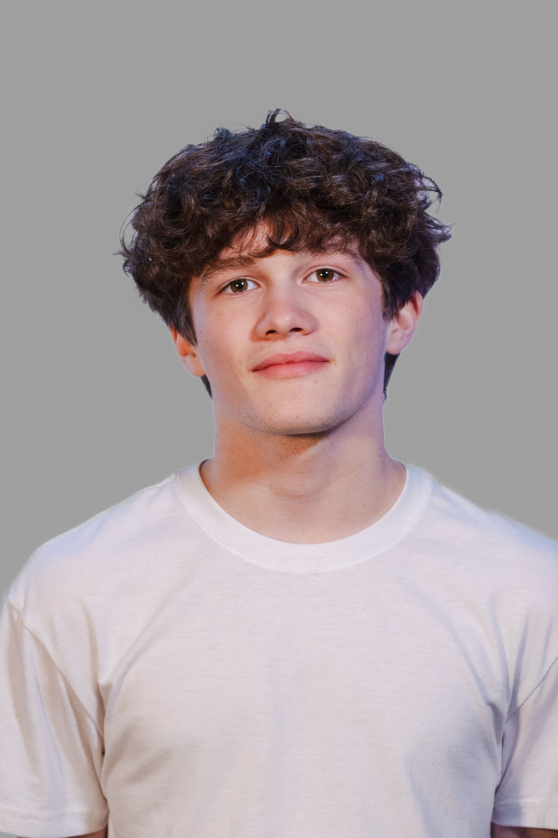Young person with curly brown hair, wearing a white t-shirt, smiling against a gray background.