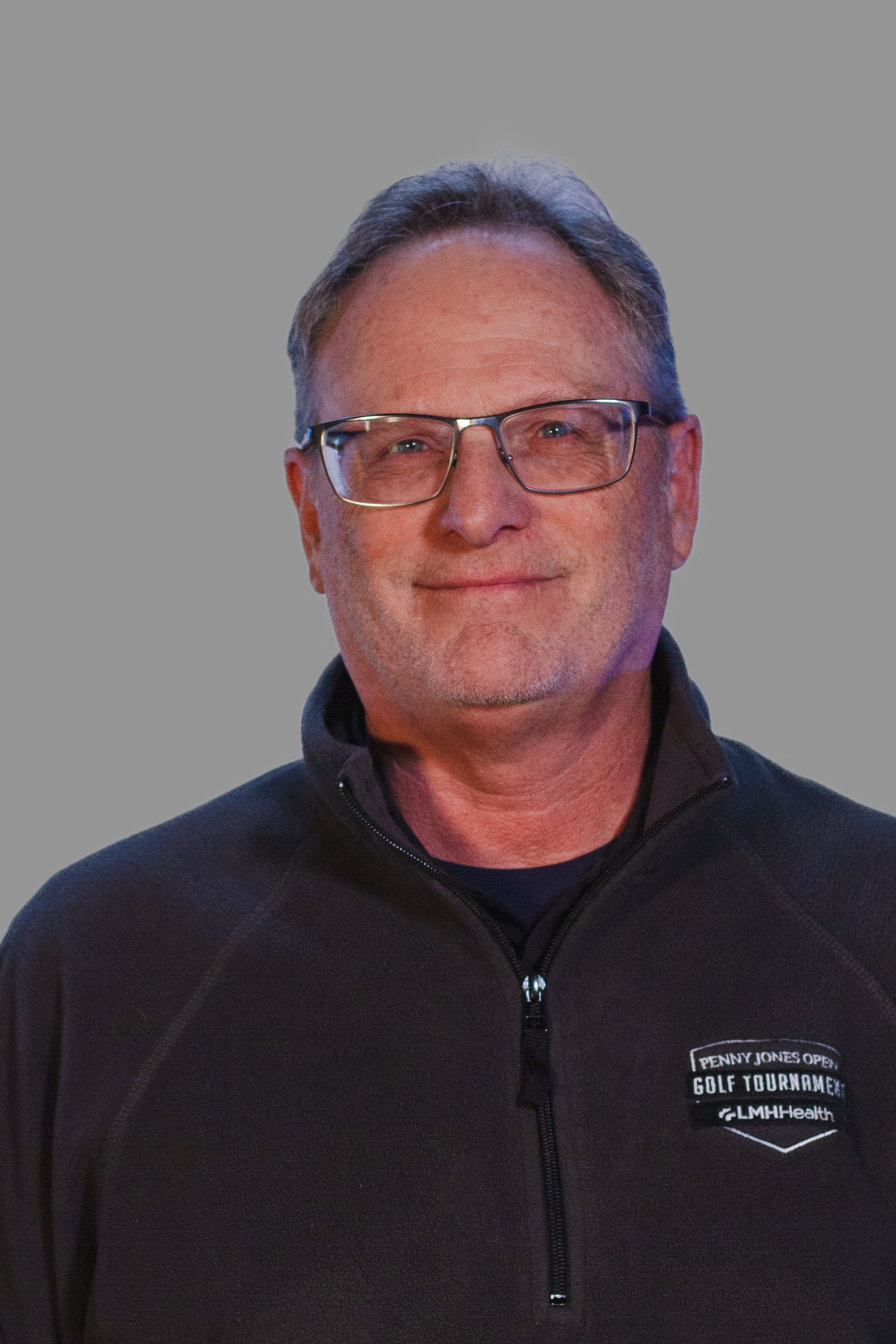 Man with glasses and short hair, wearing a dark fleece jacket, smiling. Gray background.