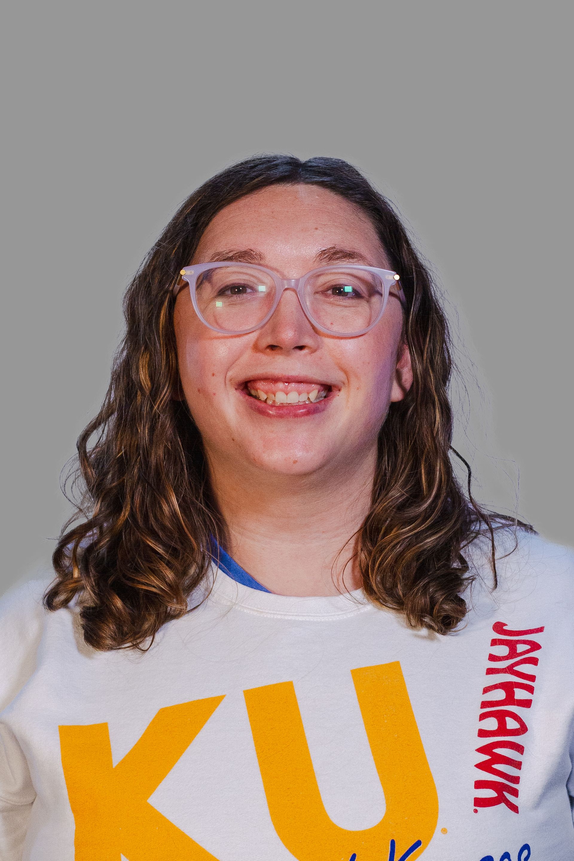 Woman smiling, wearing glasses and a KU Jayhawks shirt, with shoulder-length curly hair, against a gray background.