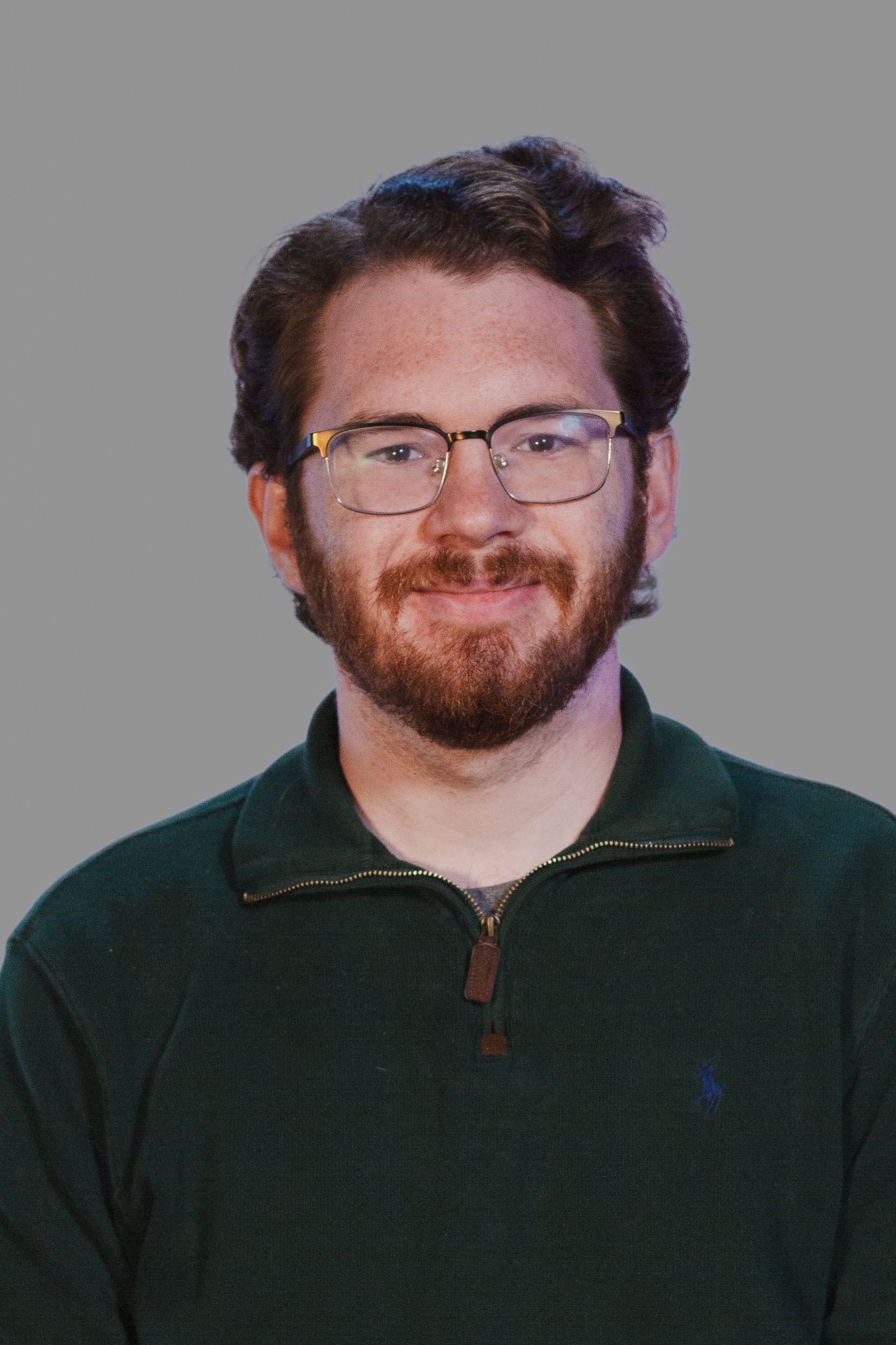 Man with glasses and beard, wearing a green sweater, smiling, against a gray background.