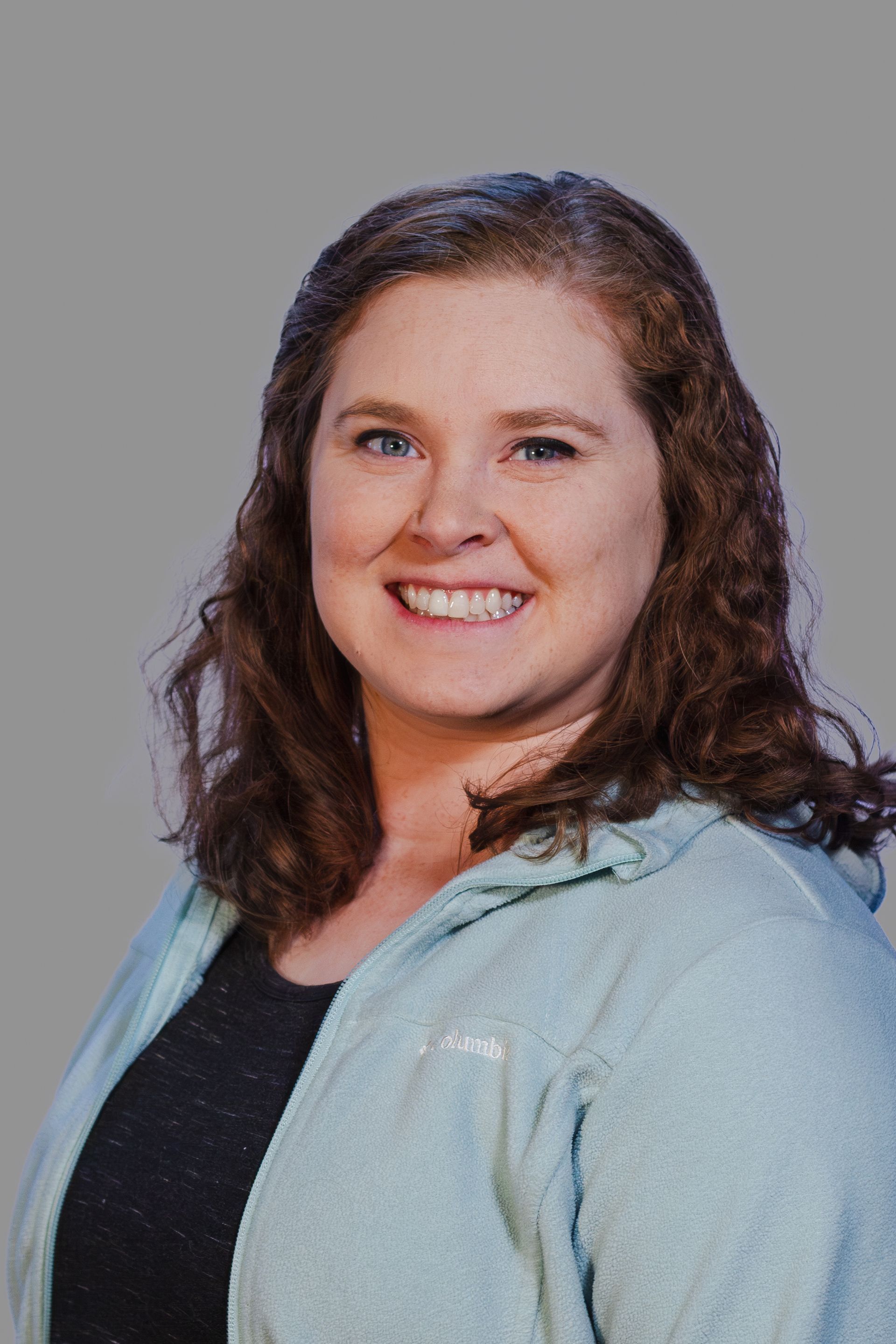 Woman smiling, wearing a light blue zip-up jacket and black top, with curly brown hair.
