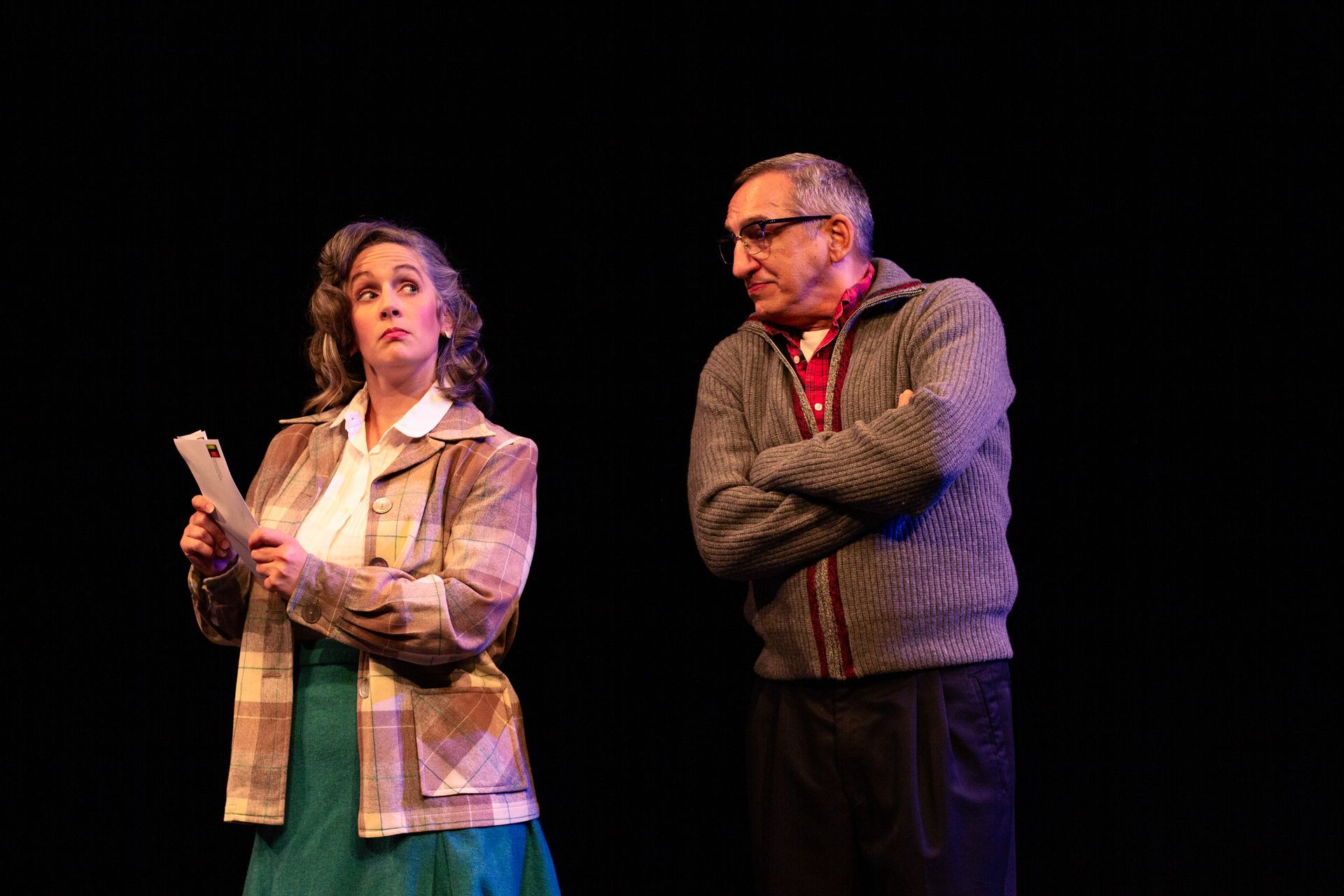 Two people on stage: a woman holding paper, looking upward, and a man with crossed arms, looking at her.