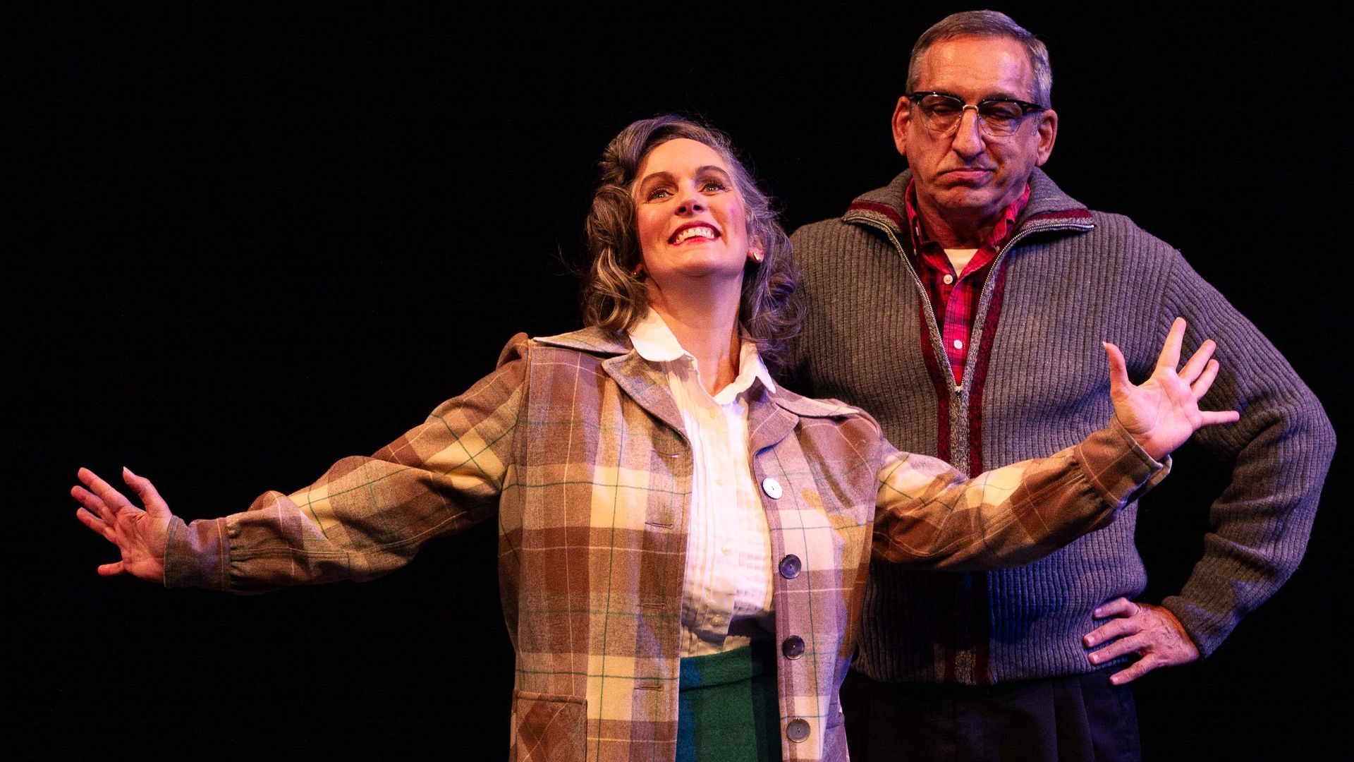 Woman with arms outstretched, man behind with hand on hip; stage setting, dark background.