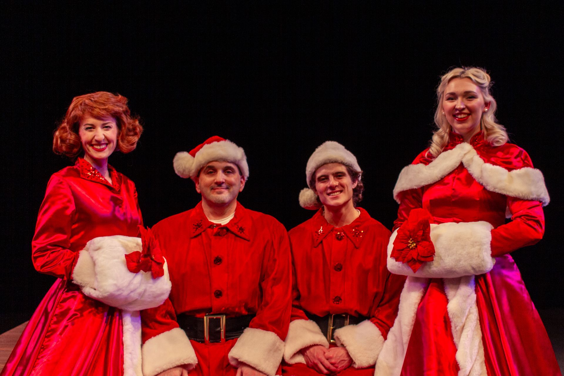 Four people in red Santa-like costumes with white fur trim, smiling against a dark backdrop.