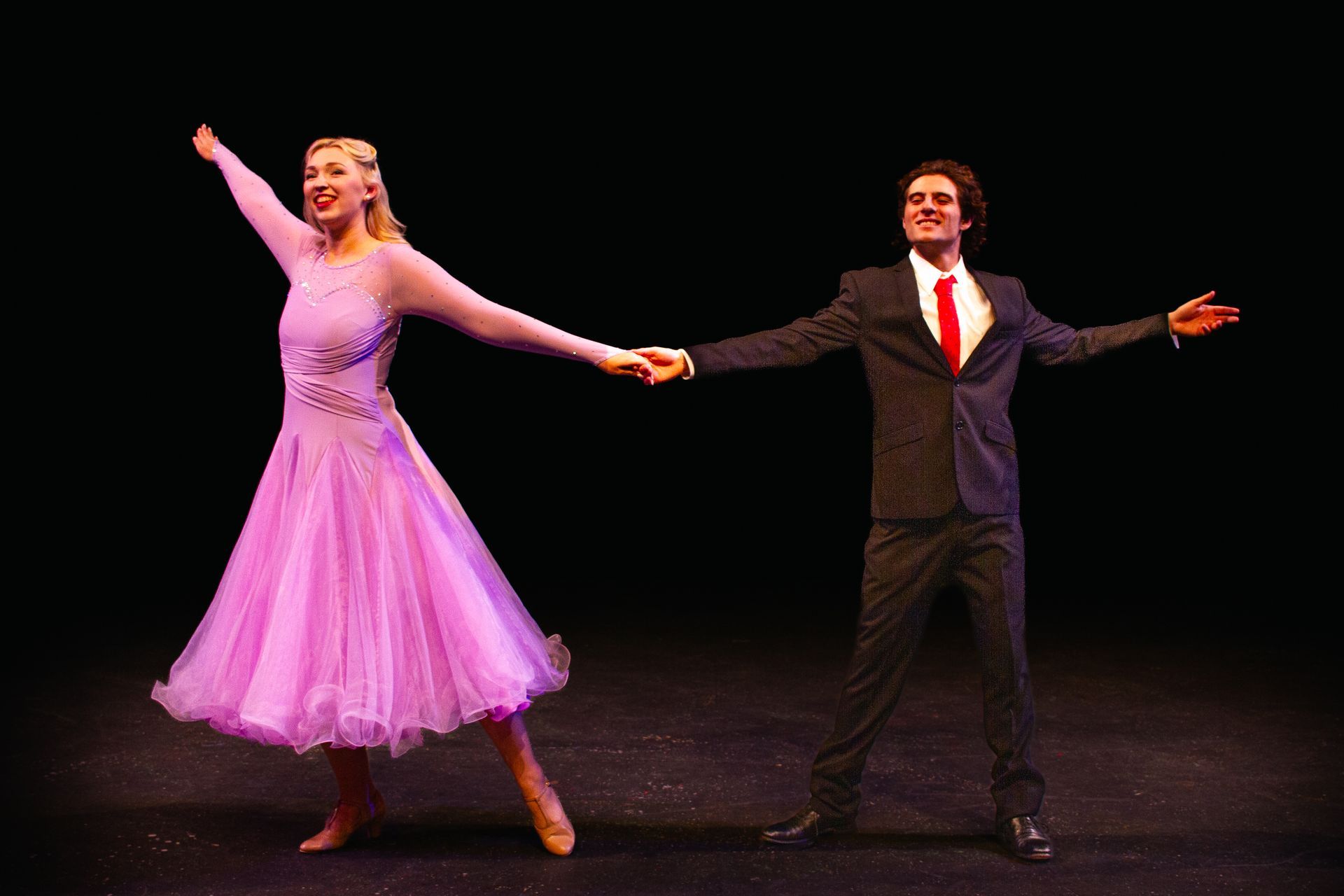 A couple dances onstage. The woman wears a lavender gown, the man a suit. Both hold hands and smile.