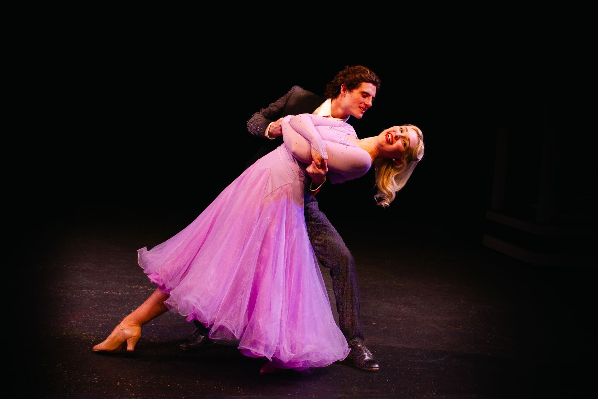 Couple dancing on a stage; man in a suit, woman in a long pink dress, both with joyful expressions.