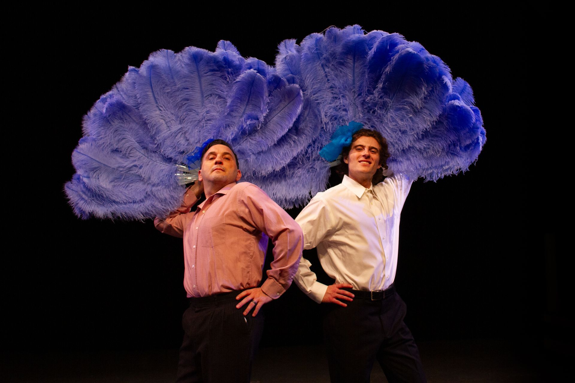Two men on a stage with large blue feather fans over their heads, wearing shirts and black pants.