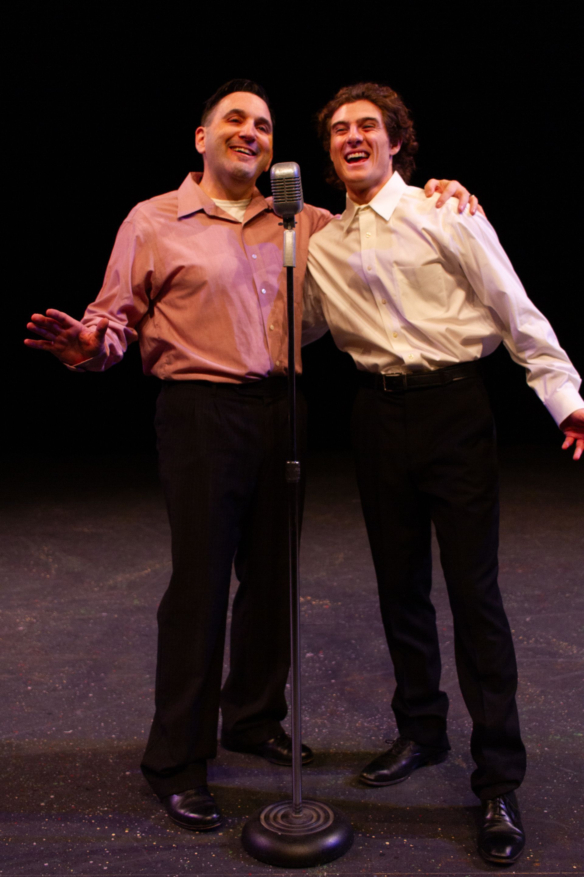 Two men, arm in arm by a microphone on a stage, smiling. One in a pink shirt, the other in white. Black backdrop.