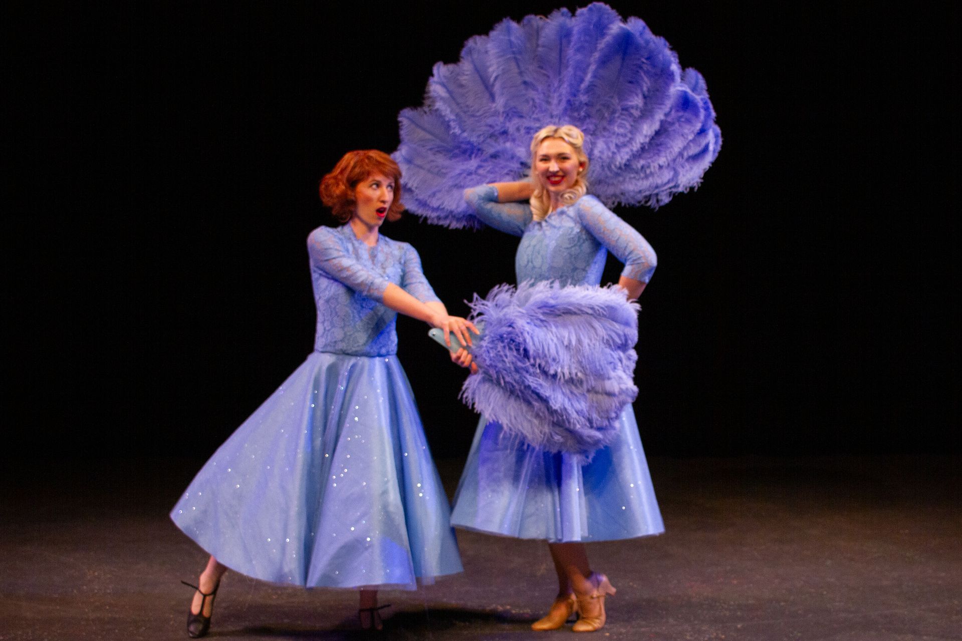 Two women in blue dresses dance on stage, holding large feather fans.