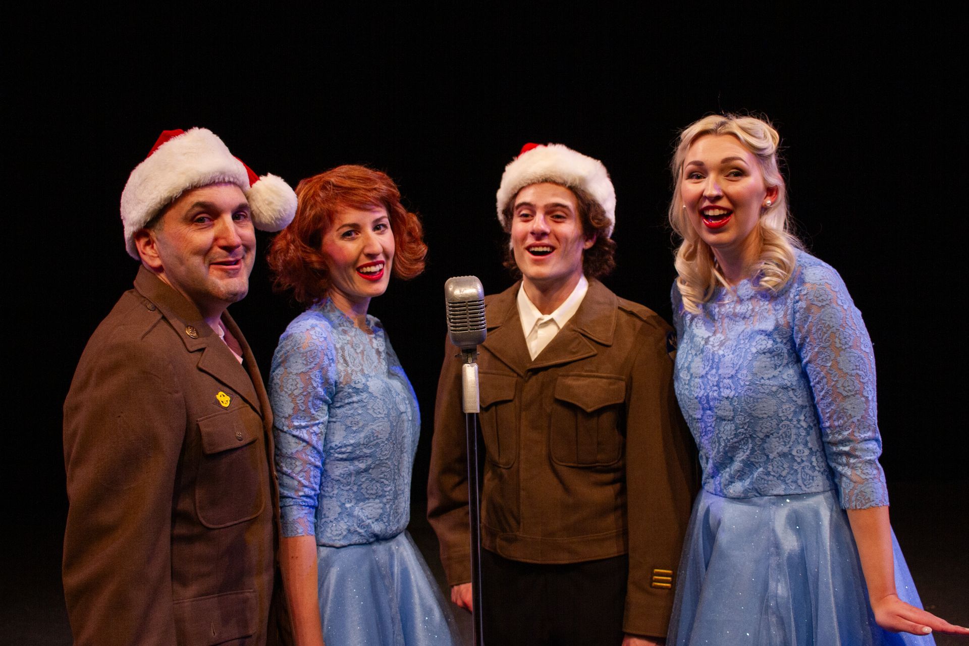 Four people in 1940s-style outfits sing at a vintage microphone, wearing Santa hats.