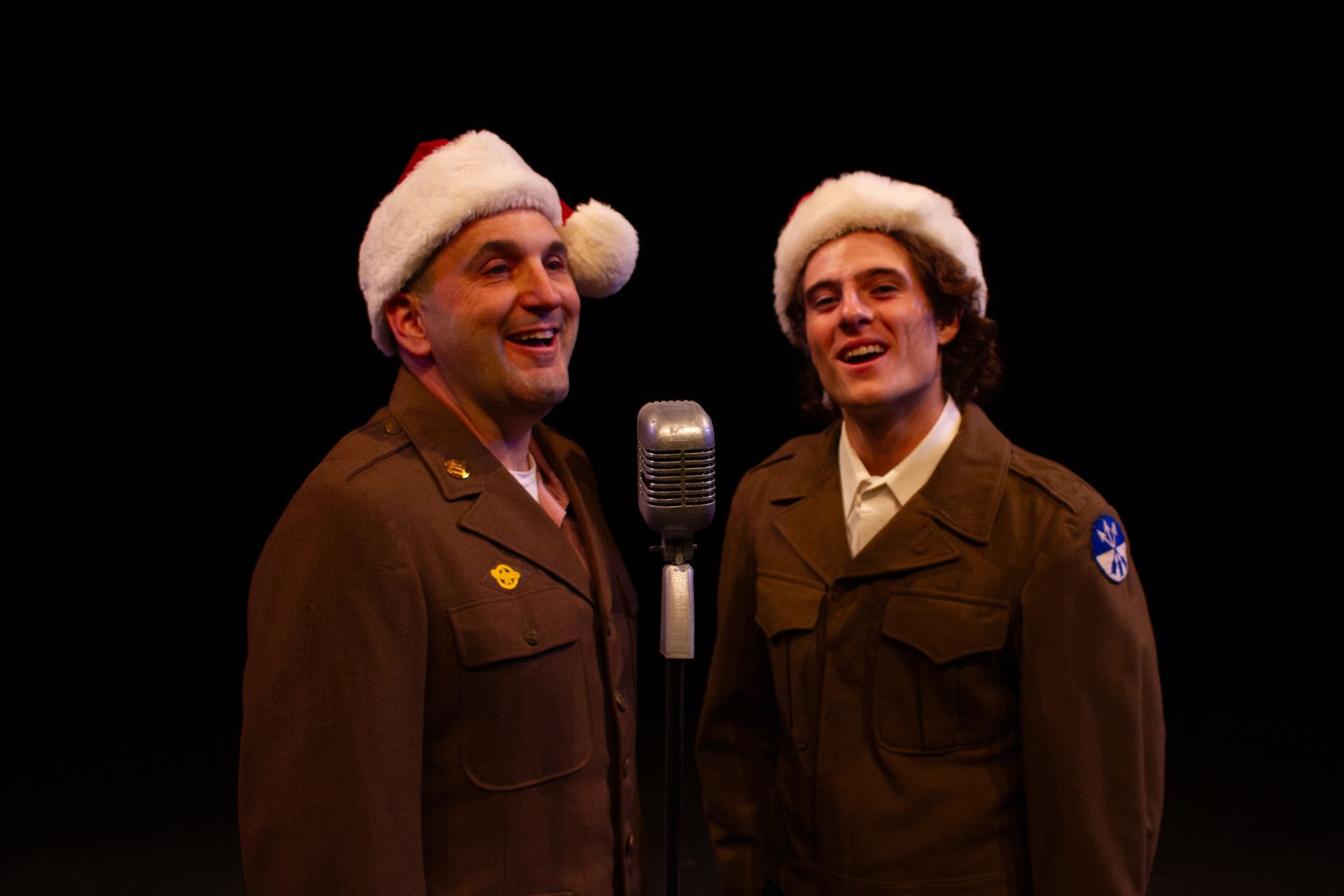 Two men in military uniforms and Santa hats sing into a vintage microphone against a black backdrop.