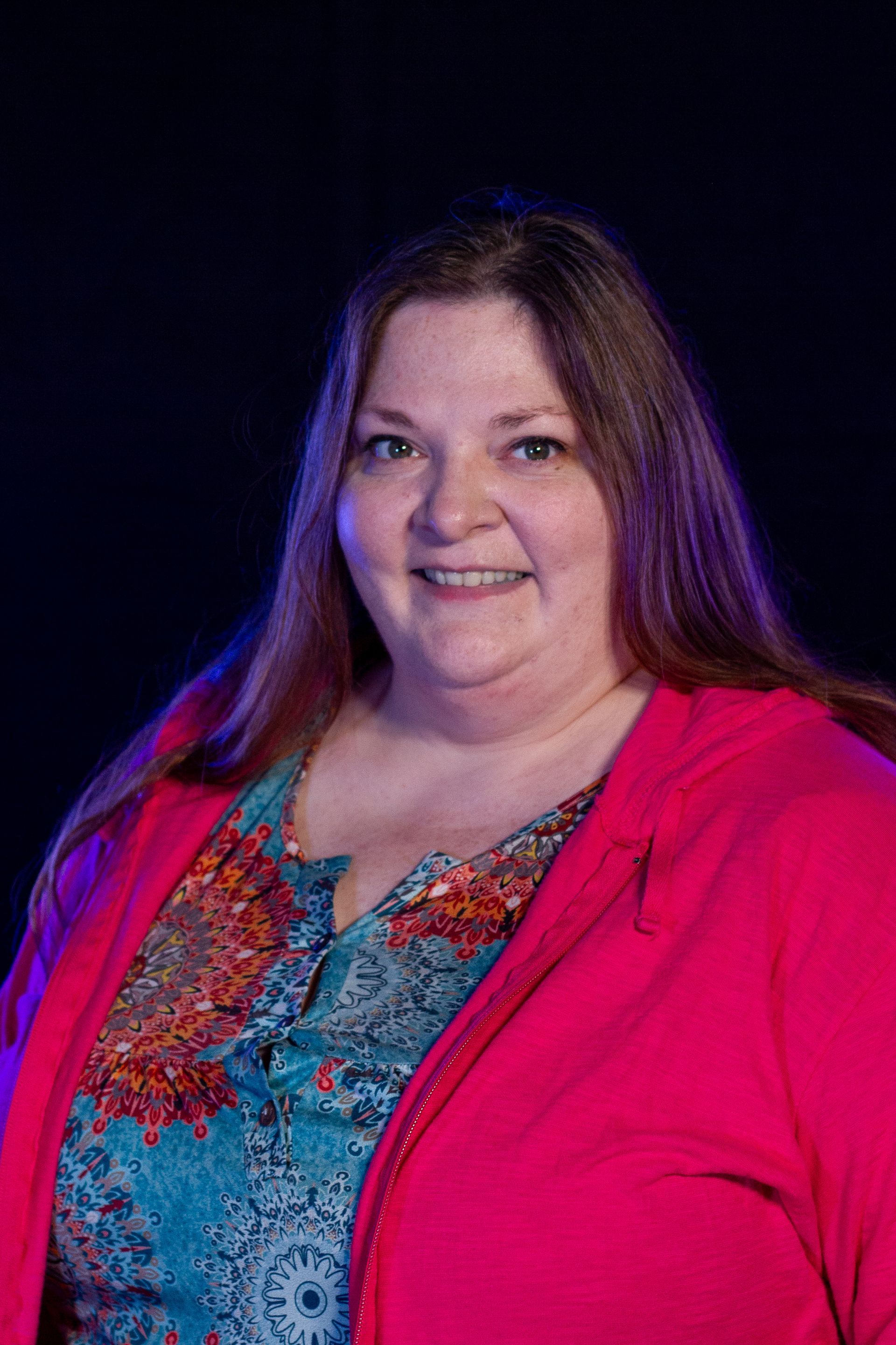 Woman with long brown hair smiles, wearing a teal patterned shirt and pink jacket against a dark blue background.