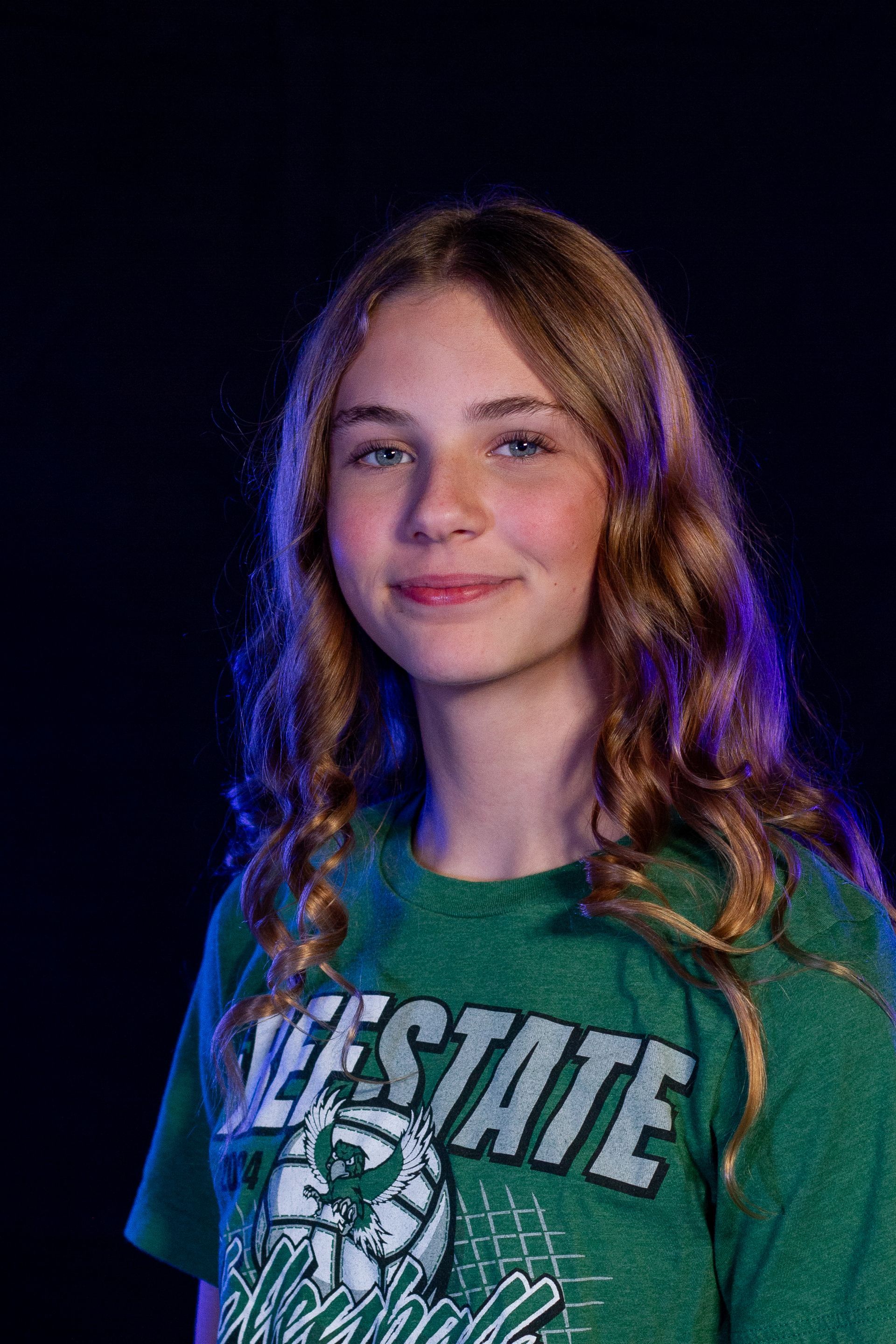 Young person with long brown hair, smiling, wearing a green shirt with a basketball graphic, dark background.