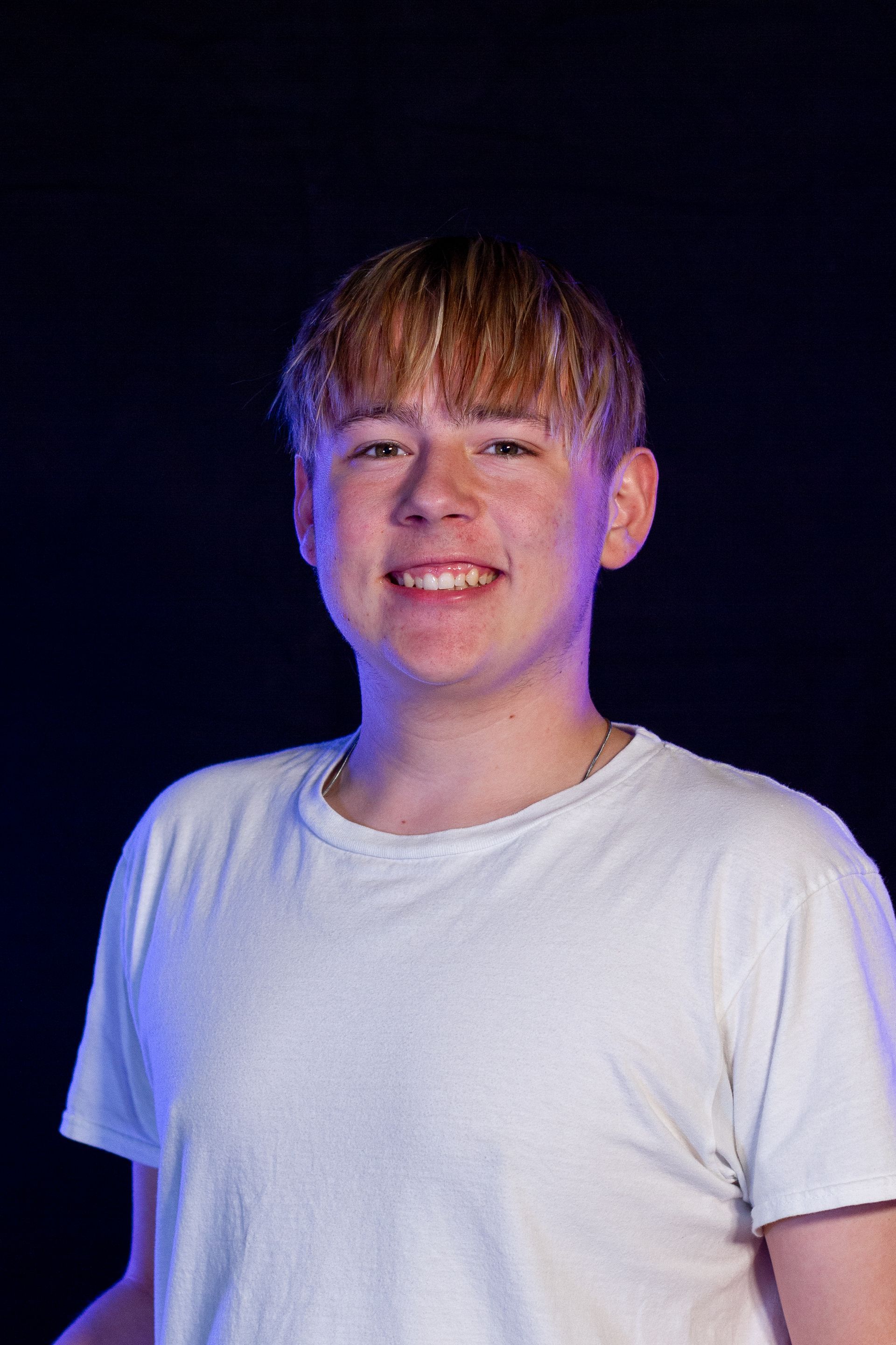Young man with a blonde fringe smiles at the camera. He wears a white t-shirt in front of a dark, blue-lit backdrop.