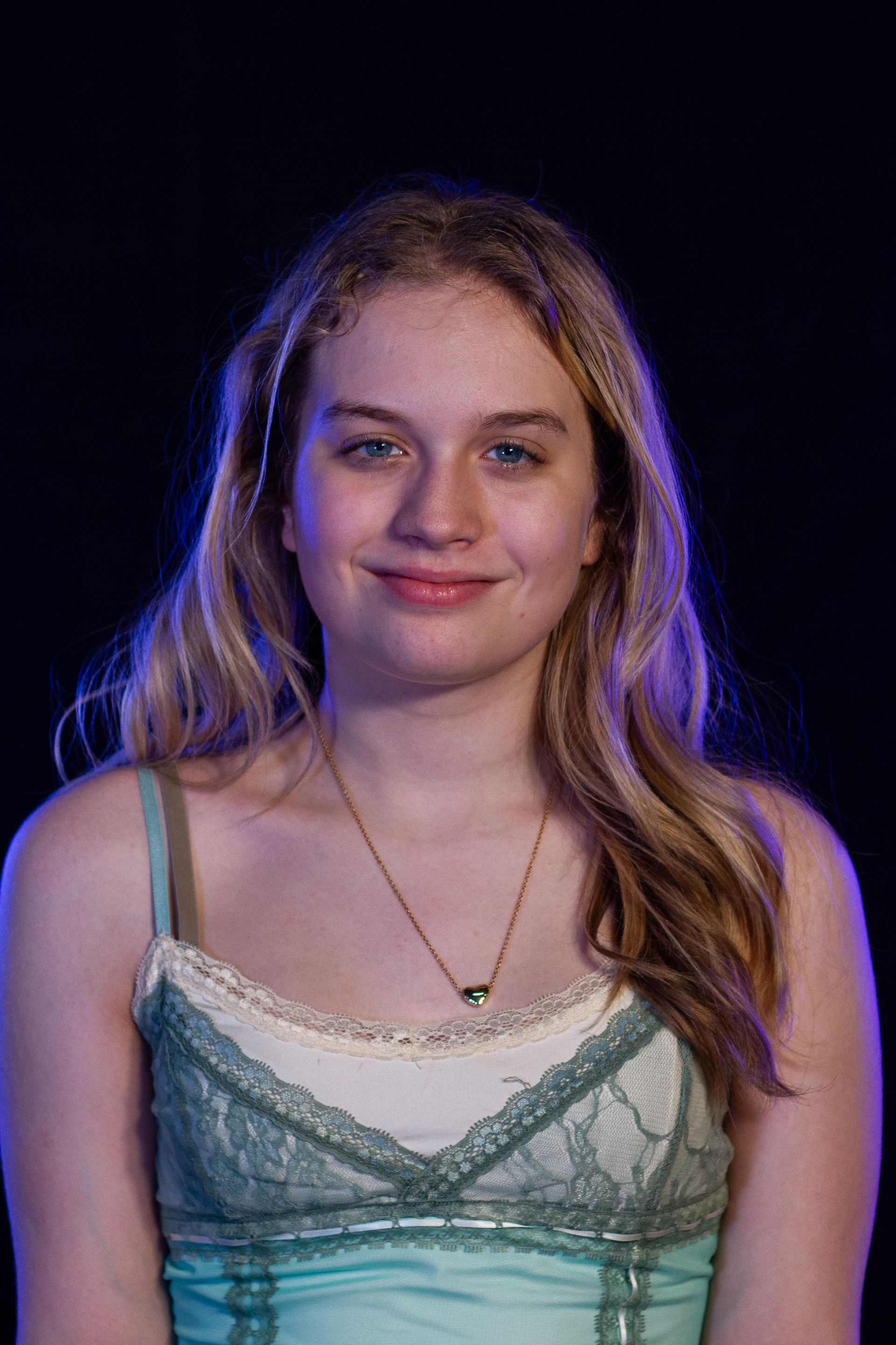 Blonde woman with blue eyes smiles at the camera, wearing a teal lace top, against a dark background.