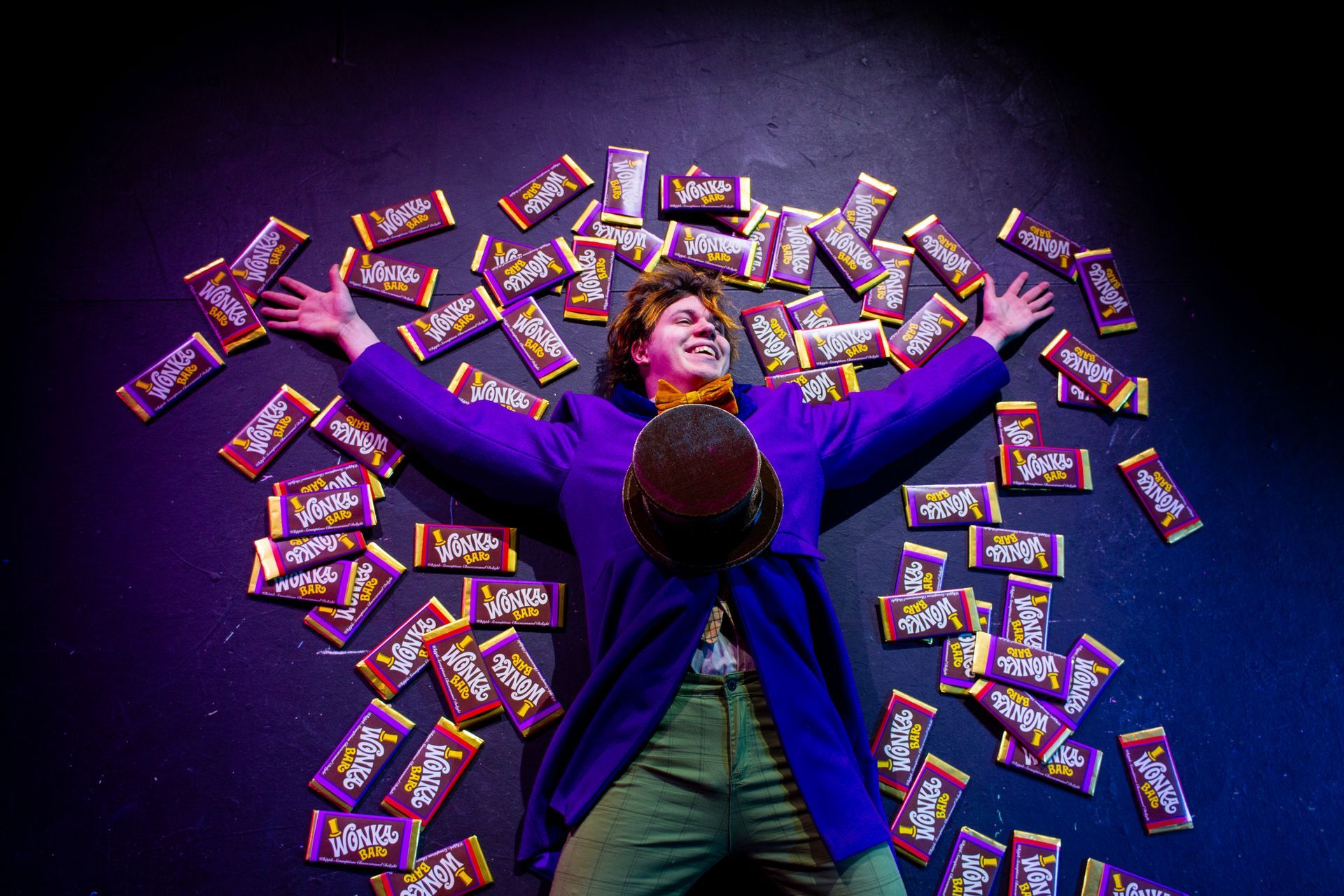 Man in purple coat, top hat, surrounded by candy bars, arms outstretched, smiling on black stage.