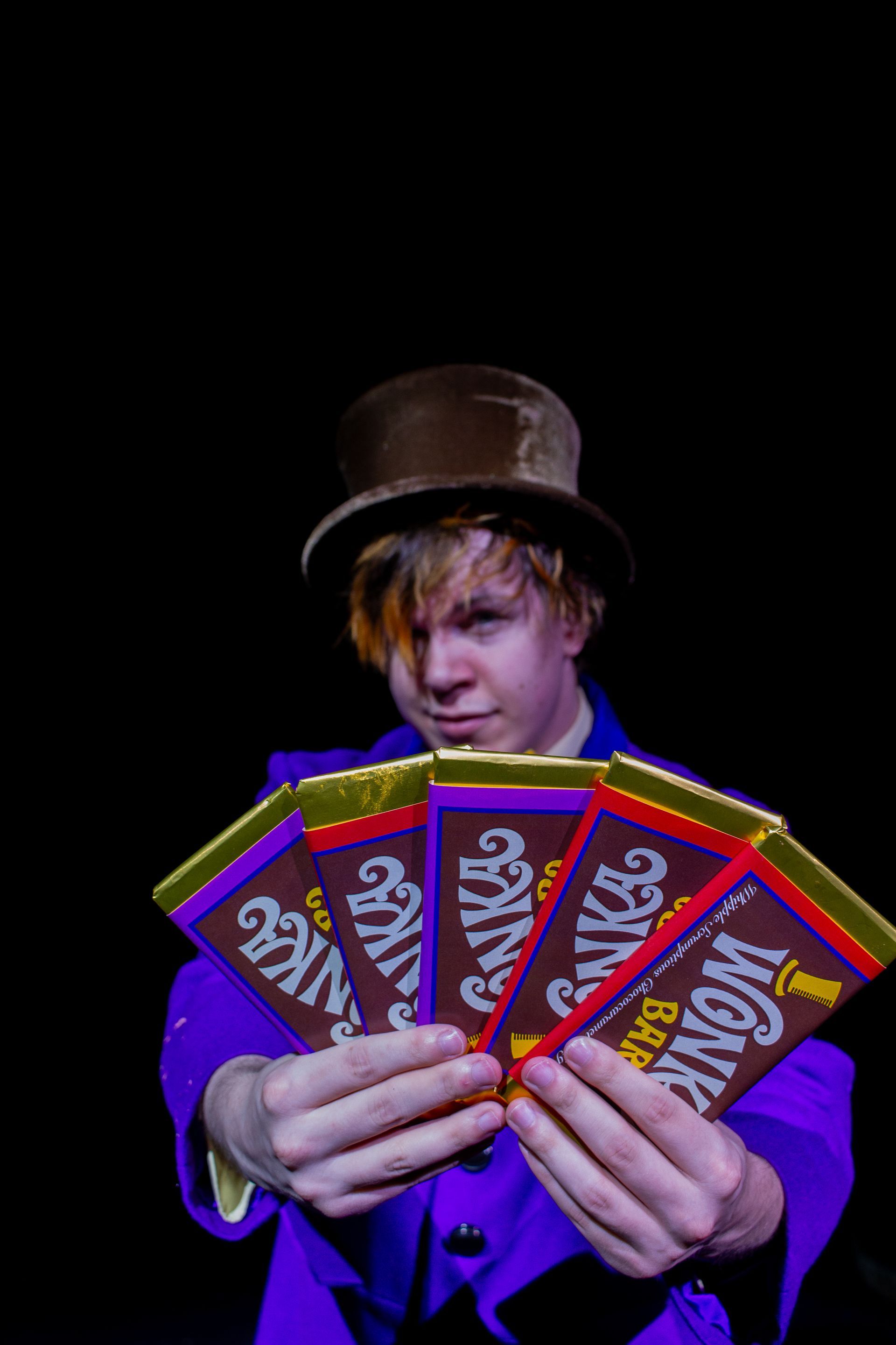 A person dressed as Willy Wonka holds up five Wonka bars against a black background.