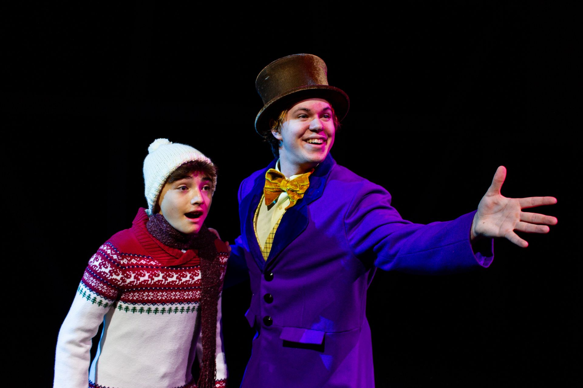 Willy Wonka in purple coat and Charlie Bucket, looking amazed, on stage with a black background.