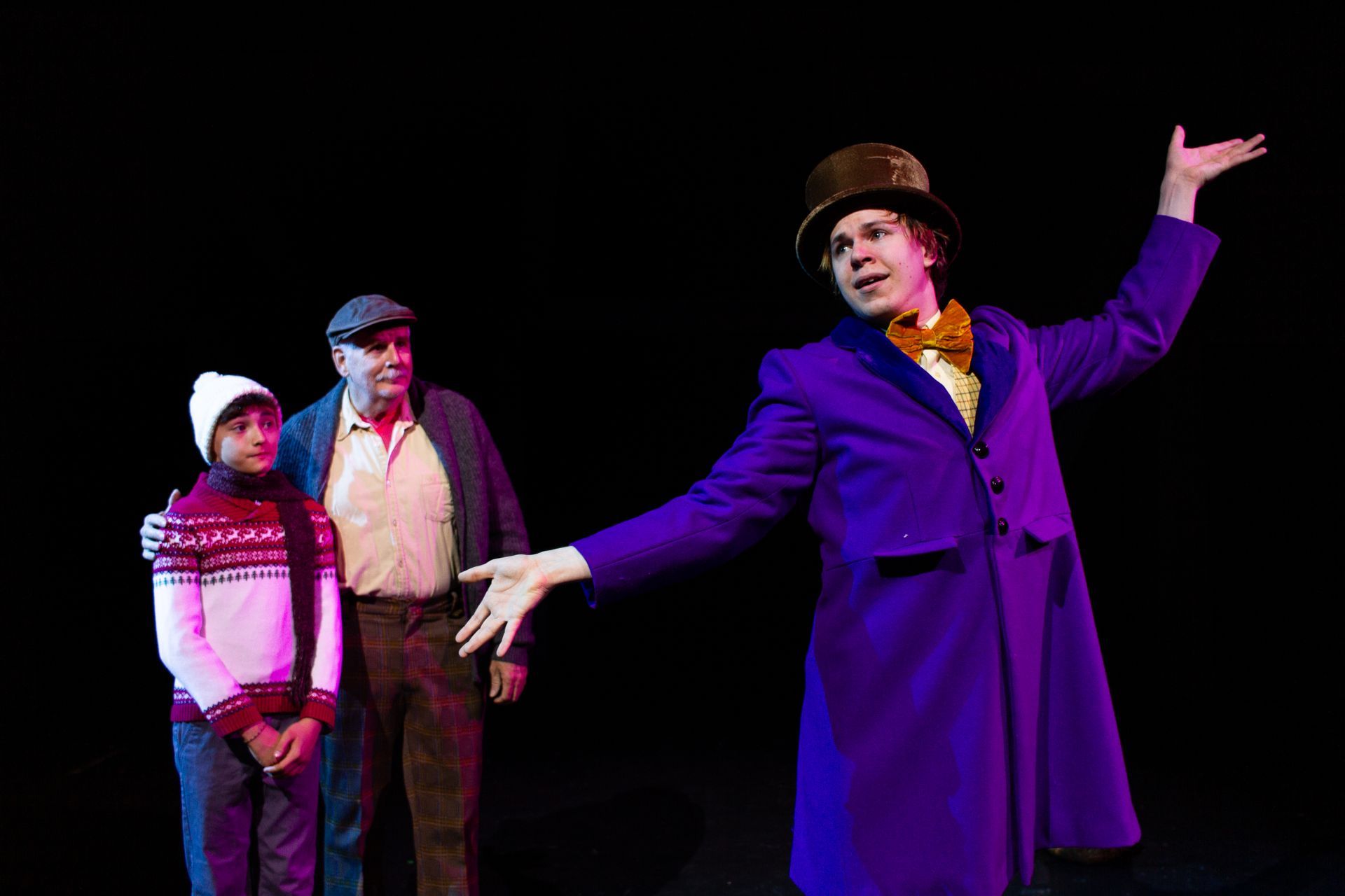 Willy Wonka in purple coat gestures, with boy and older man in the background, all on stage.