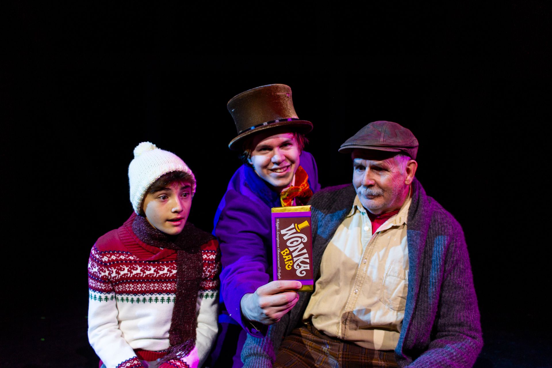 Child, Willy Wonka, and Grandpa Joe, holding a Wonka chocolate bar. Black backdrop.