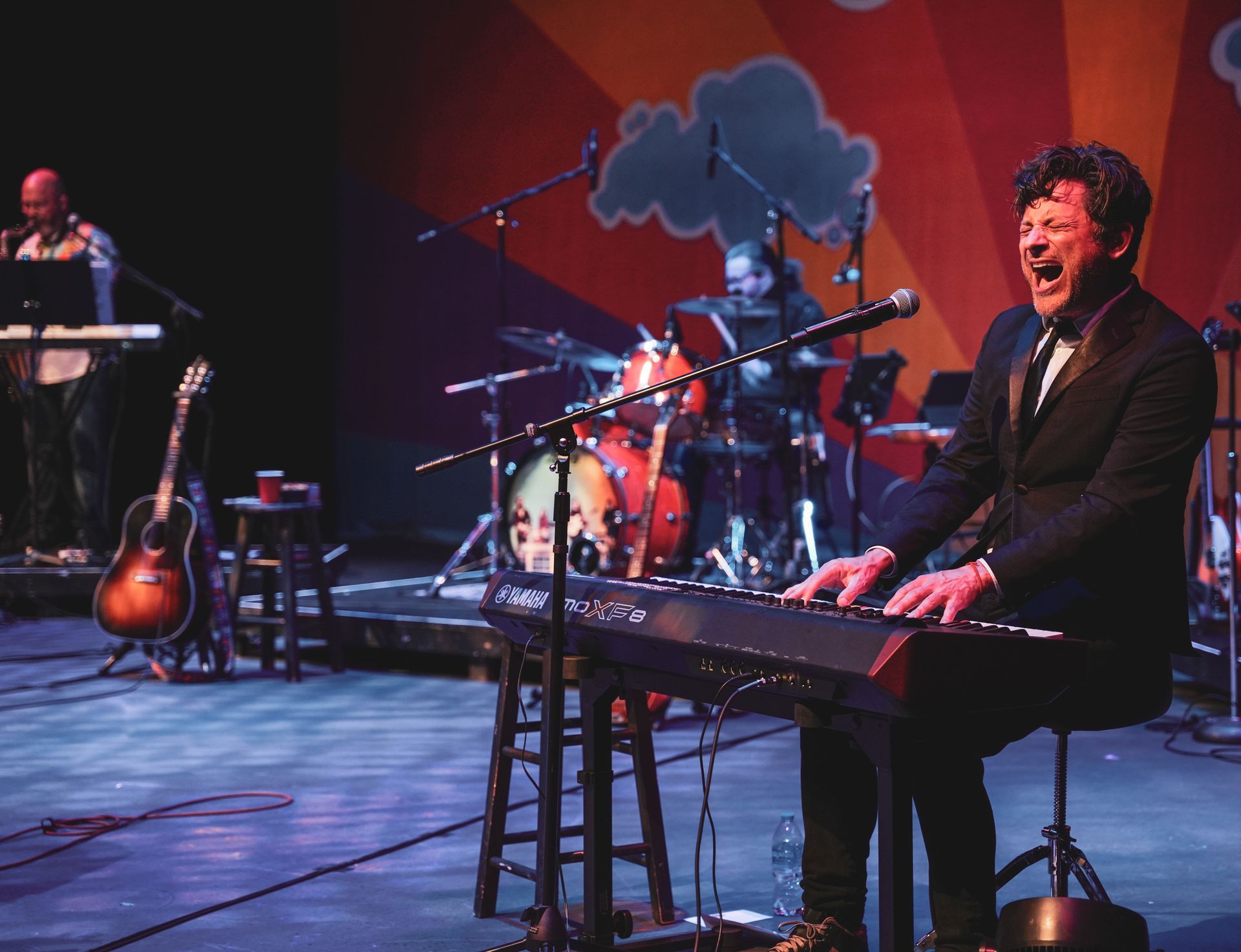 A musician playing a keyboard on stage, singing, backed by a band with drums and guitar.