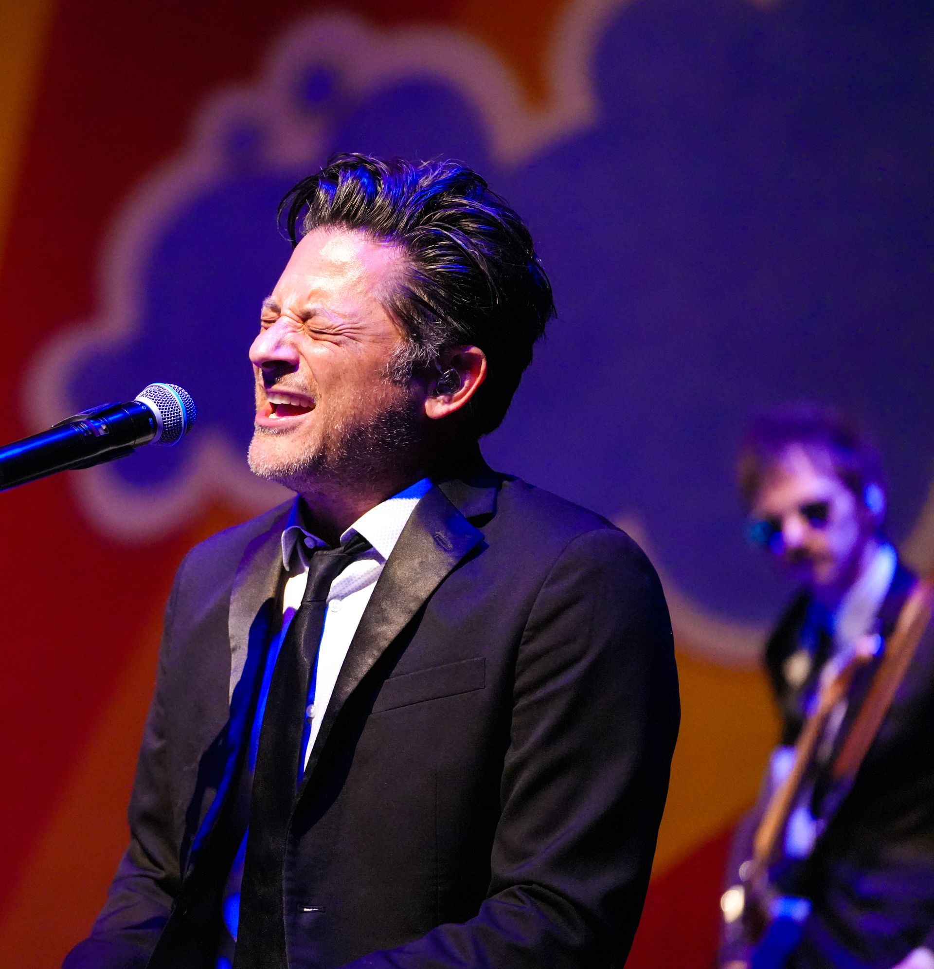 Man singing into a microphone onstage; band member in background, bright stage lights.