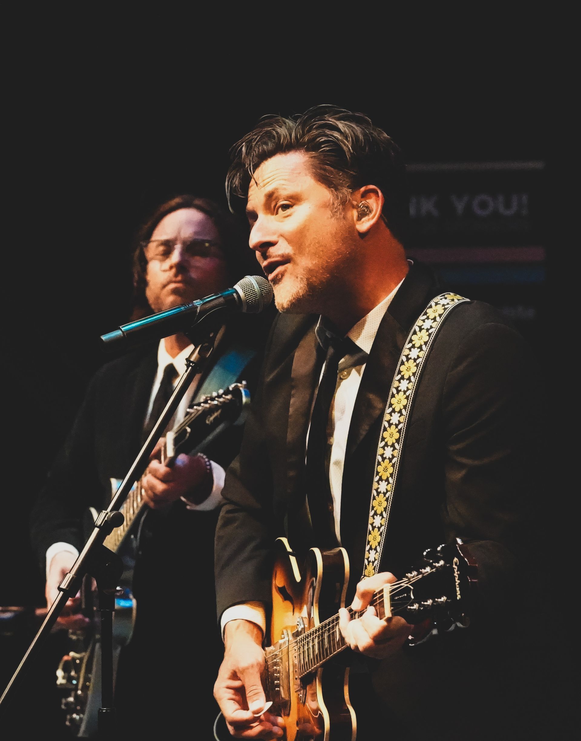 Man singing into a microphone and playing guitar onstage with a guitarist in the background.