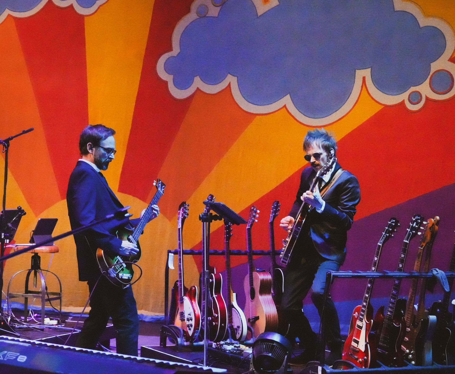 Two guitarists on stage, playing electric guitars. Colorful backdrop with sun and clouds.